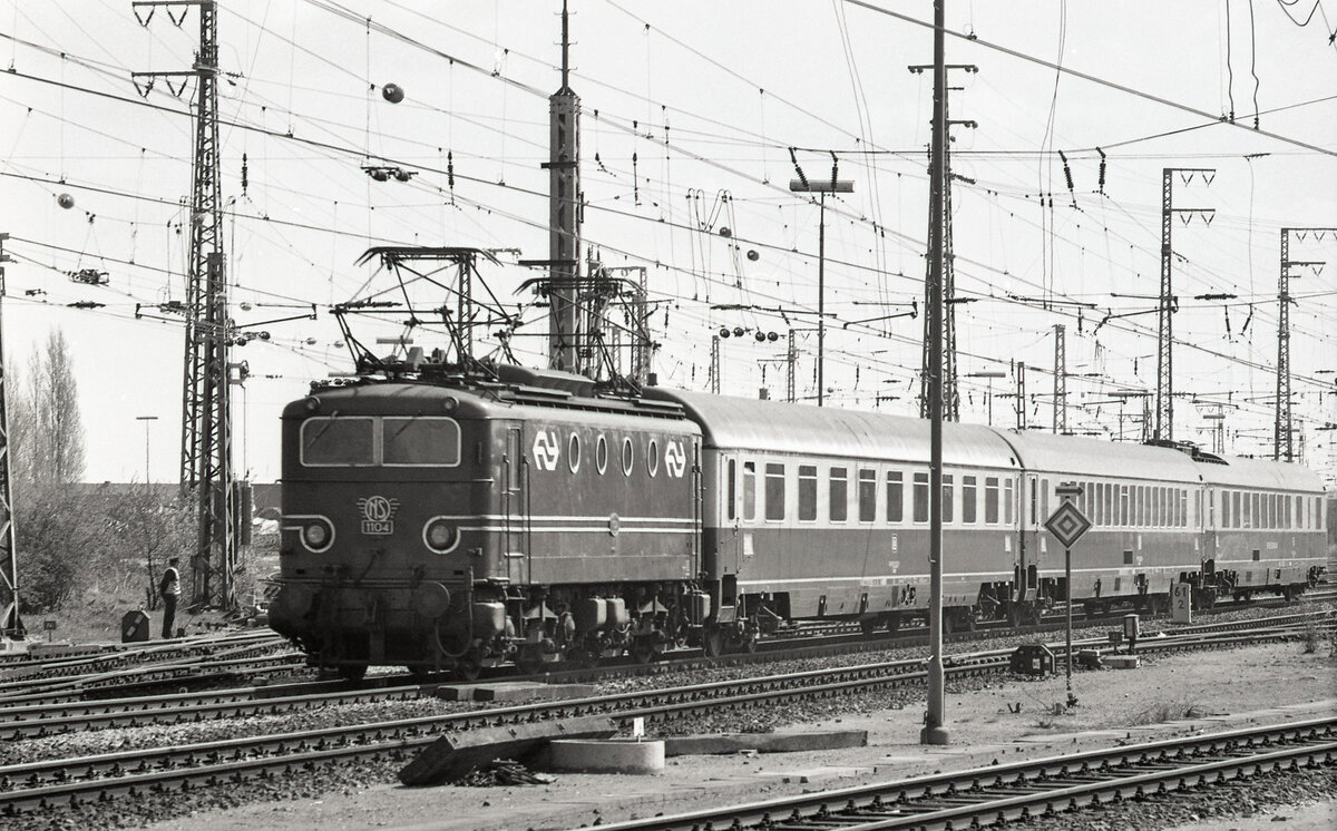 NS 1104 mit TEE-22  van Beethoven  (Frankfurt (M) Hbf - Amsterdam CS) bei der Abfahrt aus Emmerich am 22.04.1976. Wegen der Abfahrt liegen hier noch beide Stromabnehmer am Draht; wenn der Zug einmal Fahrt aufnimmt wirt der vordere Bügel gesenkt.
Planmässig sind es 1 Avm, 1 Apm und 1 WRmh der DB. Scan (Bild 90689, Ilford FP4).