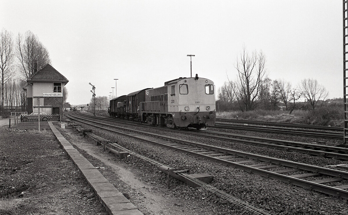 NS 2301 mit Güterzug bei der Abfahrt in Kranenburg, richtung Nijmegen, am 17.03.1978. Vermutlich Zug 46106 (Hohenbudberg - Elst). Links sieht man Stellwerk  Kw . Heute liegt hier nur noch ein Gleis für die Draisinen und ein Betonweg für die Fahrräder. Das Bahnhofsareal ist mittlerweile Urwaldartig zugewachsen. Scanbild 91375, Kodak Tri-X pan.
