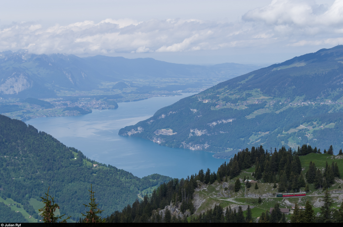 Nur ganz klein ist der Zug der Schynige Platte Bahn hoch über dem Thunersee zu sehen. Lok 63 befindet sich am 31. Mai 2015 mit ihren zwei Vorstellwagen auf Talfahrt kurz vor dem Grätli-Tunnel. Am rechten Thunerseehang befindet sich etwa auf halber Höhe das langgestreckte Dorf Beatenberg und darüber das Niederhorn mit dem Radio-Sendeturm.