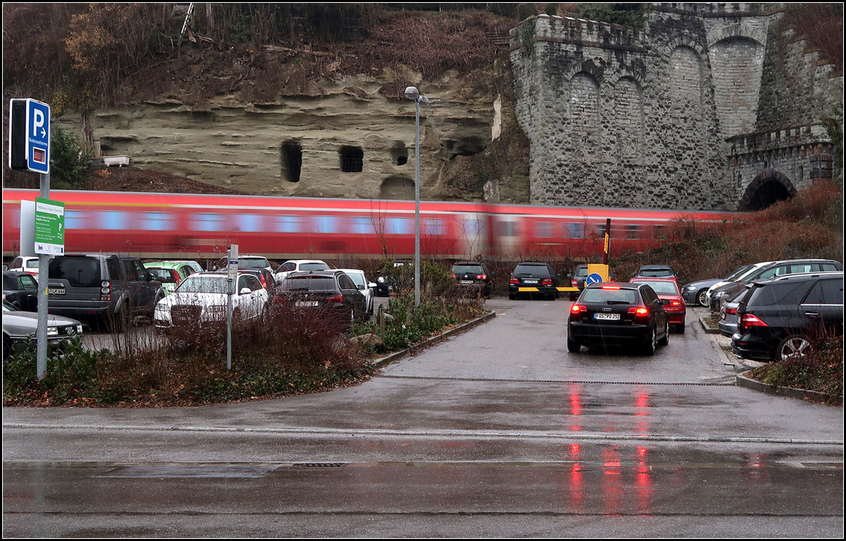 Oben hui, unten pfui -

... könnte man sagen. Neben dem Tunnelportal des Überlinger West-Tunnels befindet sich eine Felswand mit höhlenartigen Öffnungen. Darunter 'verschandelt' der Parkplatz für die Überlinger Therme das Motiv. Während ein Zug meist harmonisch in seinem Umfeld wirkt, stören Autos doch eher sehr. 
Der IRE befindet sich auf der Fahrt nach Ulm fährt in den Tunnel ein und wird am anderen Ende in Überlingen anhalten.

Die Geschichte zum Bild:
Eigentlich hatte ich während dieses Spazierganges nicht vor an diesem Portal zu fotografieren. Doch kamen wir zufällig zu einem passenden Zeitpunkt hier vorbei. Nach ein paar Bildern mit dem Regionshuttle aus anderer Perspektive wollte ich doch mal versuchen, diese Felswand mit dem IRE ins Bild zu bekommen. Mein erster Standpunkt war hinter den Autos direkt an der Strecke. Auch da war der Plan mit Bewegungsunschärfe zu arbeiten. Da aber ein Schneeschauer einsetzte suchte ich kurzerhand nach einer Unterstellmöglichkeit, die in Form eines Bus-Wartehäuschen auch zu finden war. Dann war es noch spannend ob zum Zeitpunkt der Zugdurchfahrt Fußgänger oder ein LKW bzw. Bus die Bahn verdecken würden, was dann zum Glück nicht der Fall war. Ein zufälliges Plus ist dann noch die Spiegelung der roten Rücklichter auf dem regennassen Asphalt, der einen vertikalen Kontrapunkt zum horizontalen roten Streifen des Zuges setzt...

11.02.2017 (M)

