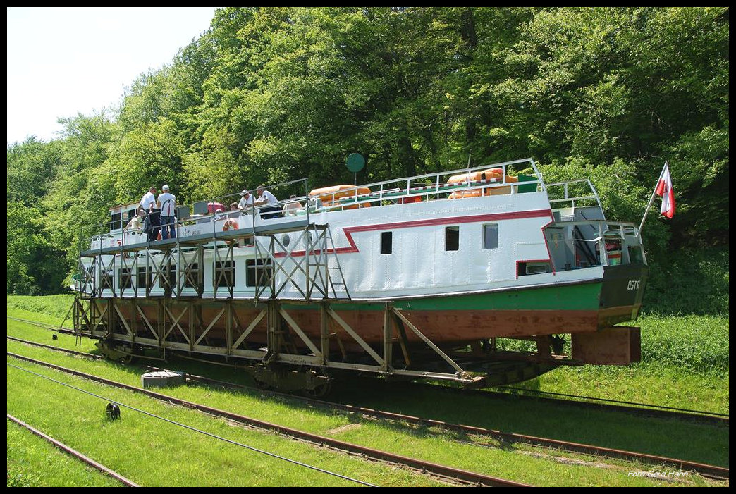 Oberlandkanal bei Buczyniec am 22.05.2012: Der Oberlandkanal in Polen ist heute  eine viel besuchte touristische Attraktion; denn dort wird man als Passagier auf dem Schiff mittels einer Standseilbahn über fünf Berge gezogen. Eine dieser Bergpassagen befindet sich bei Buczyniec. Am 22.05.2012 hielt ich eine solche Fahrt im Bild fest: Hier hat der Rollwagen mit der  MARABU  die Bergspitze überschritten und rollt nun talwärts in Richtung Ostroda. Deutlich kann man die vier Achsen des Rollwagen erkennen. 