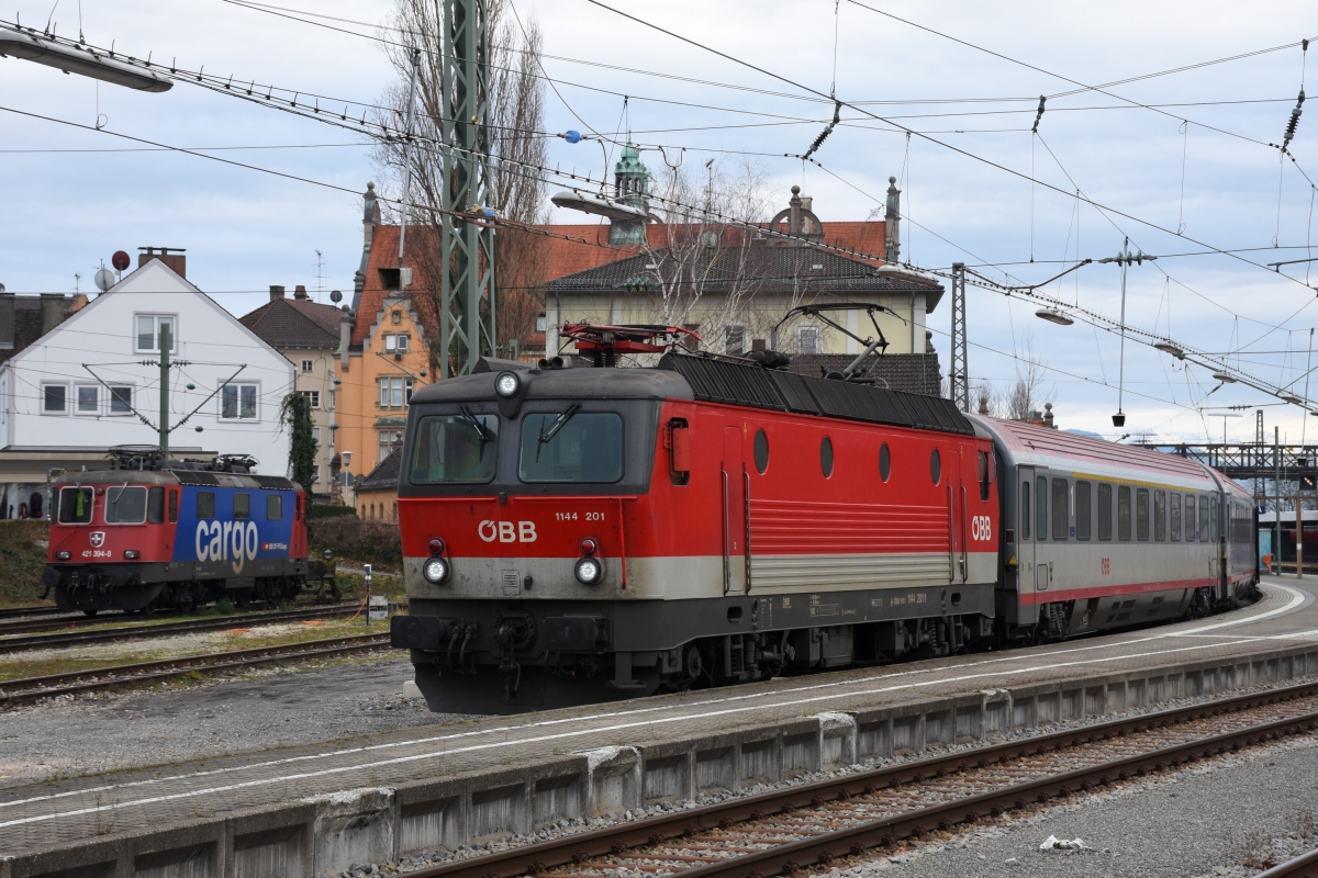 ÖBB 1144.201 übernimmt in Lindau den von Münster kommenden IC 119 zur Weiterfahrt nach Innsbruck. Im Hintergrund hat SBB CARGO 421 394 auf einem Nebengleis noch knapp 3 Stunden zu warten, bevor sie sie EC 192 von München nach Zürich übernehmen darf (28.01.2018).