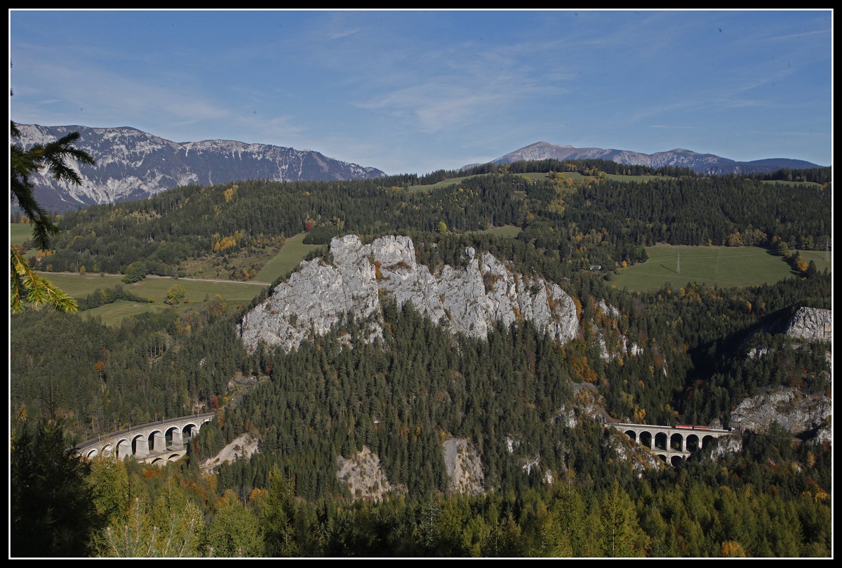 Panoramabild der Semmeringbahn mit Kalter Rinne , Poleroswand und Krauselklause. Die Berge im Hintergrund sind die Rax und der Schneeberg. Der Lokzug der auf dem Krauselklauseviadukt fährt, findet wegen seiner Winzigkeit kaum Beachtung.