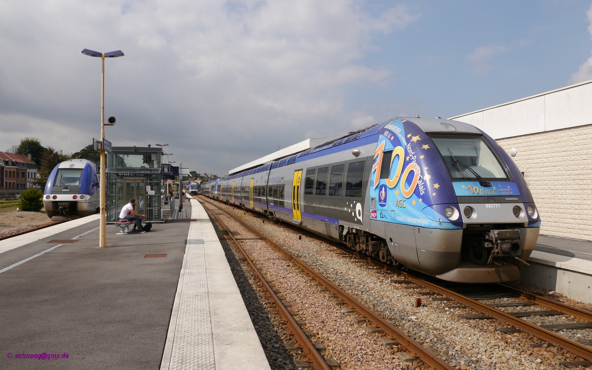 Provinzbahnhof mit großem AGC-Treffen.
Die Triebzüge des Typs AGC (Autorail à Grande Capacité  „Triebwagen mit großer Kapazität“) wurden von Bombardier hergestellt.
Es gibt sie in verschiedenen Varianten als Diesel- (XGC), Elektro- (ZGC), sowie als Hybrid-Triebzüge für Diesel und Elektrobetrieb (BGC).
Die Hybridzüge gibt es in der Variante für Diesel und Gleichspannung 1,5 kV (Bimodal Bi), sowie in der Variante für Diesel und Gleichspannung 1,5 kV und Wechselspannung 25 kV (Bimodal-Bicourant BiBi).
Von 2004-2011 wurden insgesamt 700 dieser Triebzüge in den unterschiedlichen Variaten ausgeliefert.
Im Juni 2011 wurde der 700. und damit Letzte an die Region Nord-Pas-de-Calais ausgeliefert.
Der letzte dieser ganz im Norden Frankreichs im Bombardier-Werk in Crespin in der Region Nord-Pas-de-Calais gebauten Züge wurde mit einer entsprechenden Werbung versehen.
Es ist der rechts im Bild zu sehende SNCF-B82771, an noch der X76711 gekoppelt ist.
In der Bildmitte fährt gerade der SNCF-X76613 auf dem Weg nach Arras ein, während links der Kopf des SNCF-B82737 zu sehen ist, der als TER42321 von Arras nach Le Touquet fährt.

2014-07-26 Saint-Pol-sur-Ternoise