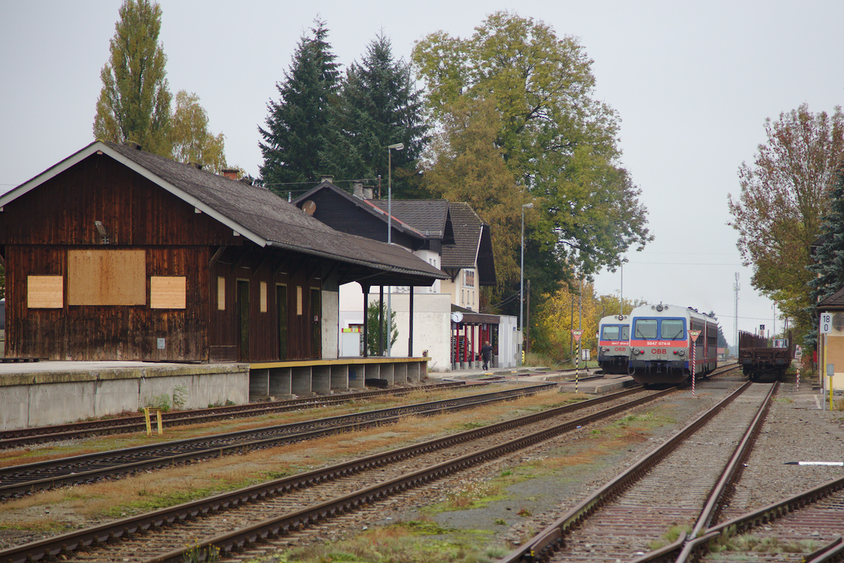 Recht stilvoll präsentiert sich der Bahnhof Mattighofen an einem trüben Herbsttag. Lediglich zwei Handymasten, einer davon ganz rechts stören das ansonsten schöne Ensemble.
Um 10:30 kreuzen hier täglich der Zug nach Braunau mit dem nach Salzburg Hbf. Am linken Kreis warten der 5047 064 und der 5047 084, wobei letzterer durch den gerade einfahrenden Zug nach Braunau am rechten Gleis verdeckt wird. Dieser bestand aus dem 5047 071 und 5047 074. (24.10.2015)
