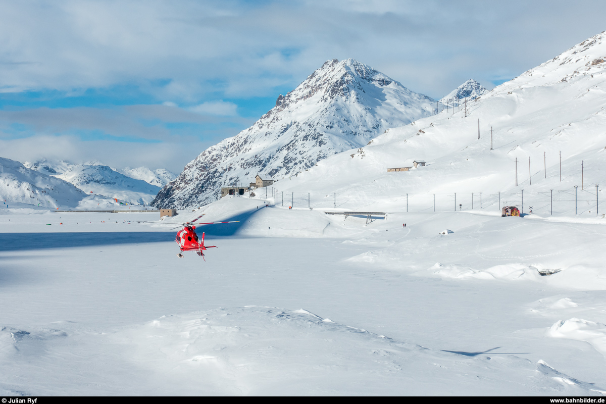 RhB Dampfschneeschleuderfahrt am 28. Januar 2018. <br>
Während der Helikopter der Schweizerischen Rettungsflugwacht Rega zur Landung ansetzt, wartet die Dampfschneeschleuder Xrotd 9213, geschoben vom Bernina-Krokodil Ge 4/4 182, beim Bahnübergang auf ihren Einsatz bei der dritten Schleuderdemo des Tages.