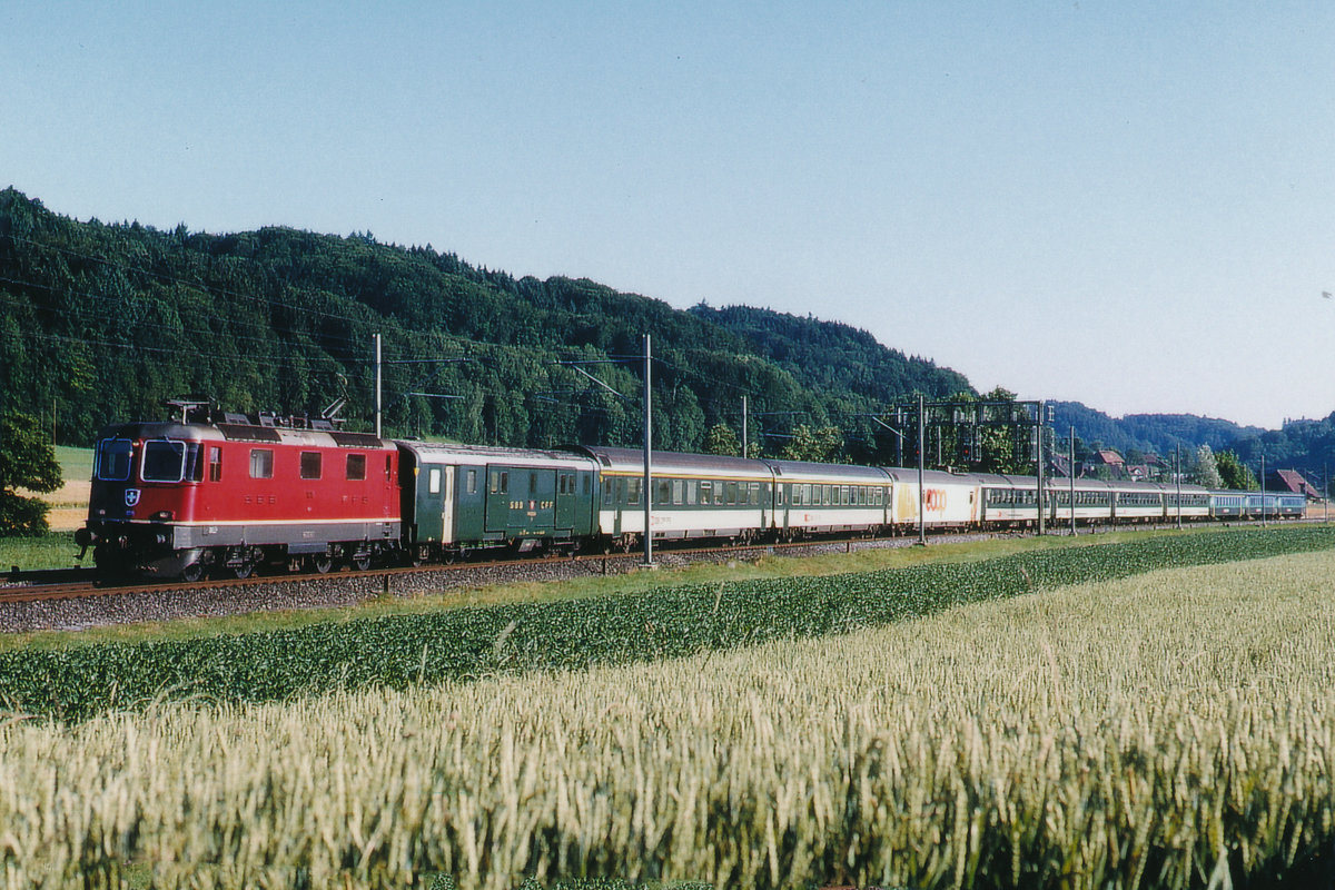 SBB: Re 4/4 II im Personenverkehr.
Schnellzug Genève-St. Gallen mit einer nicht erkennbaren Re 4/4 II der zweiten Serie bei Bollodingen unterwegs im Juli 1999.
Besondere Beachtung gilt nebst der bunten Anhängelast dem Gepäckwagen sowie dem Coop Shop-Wagen.
Foto: Walter Ruetsch 