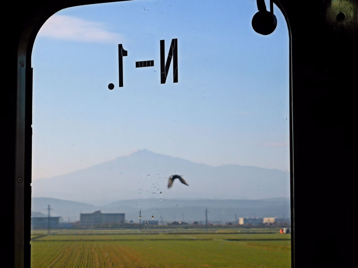 Serie 701 des Bezirks Akita - auf der Nordwest-Hauptlinie: der Nordteil dieser Strecke wird durch den riesigen, 2236 Meter hohen Vulkan Chôkaisan dominiert. Im Morgendunst nähert sich Zug 701-1 (bezirksintern als Zug N-1 gekennzeichnet) von Akita her dem Fuss des Vulkans. Bei Ugo Iwaya, 2.November 2013.  
