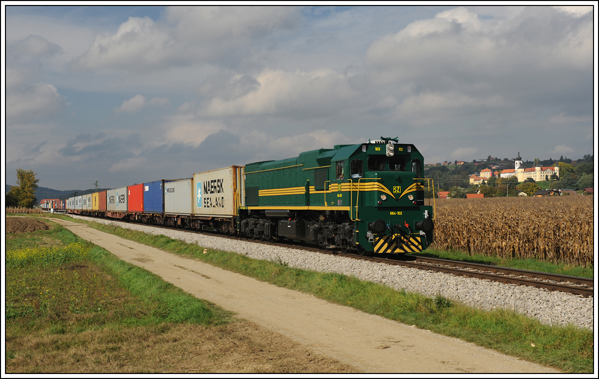 Sowohl am 8. als auch am 9.10.2013 war ich an dieser Stelle, aber niemals hatte ich Glck. Am 10.10.2013 klappte dann alles. Ich positionierte mich strategisch gnstig nchst Velika Nedelja um a) Zge Richtung Sden (Pragersko) fotografieren zu knnen und b) Zge Richtung Osten (Hodo) bereits aus der Ferne sehen zu knnen. So war es dann auch – in der Ferne sah ich 664-102, welche mit einem Metrans Containerzug Richtung Hodo fuhr, und berstellte an diese hier gezeigte Fotostelle kurz nach der Haltestelle Velika Nedelja. Im Hintergrund ist die Pfarrkirche der hl. Dreifaltigkeit und die Burg des Deutschritterordens, in der heute eine Volkskundliche Sammlung untergebracht ist, zu sehen.