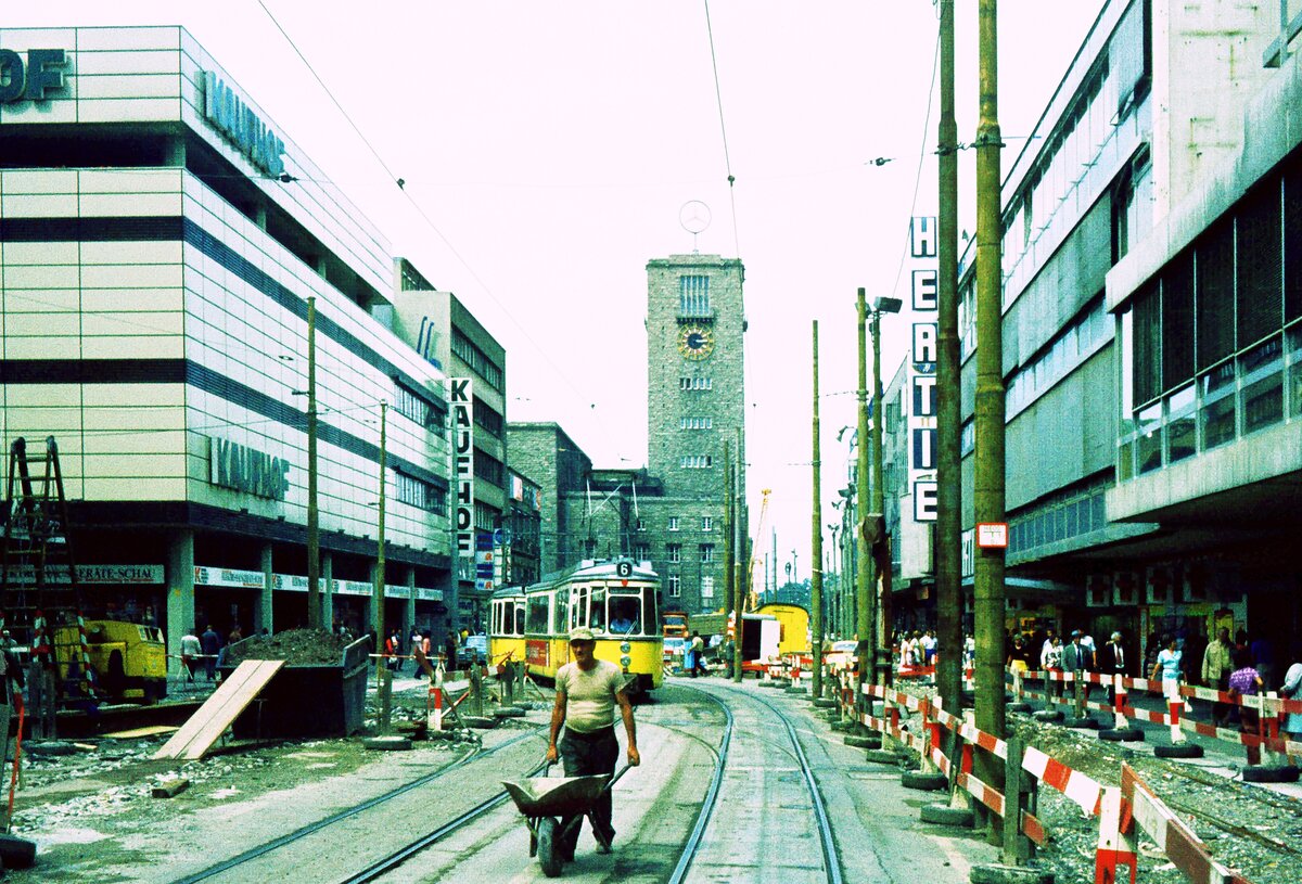 SSB Stuttgart__Gleis- und Tunnelbau__Der Mann mit der Schubkarre gibt den Takt vor ? ( Quietsch quietsch... ) Naja, es ist ja Platz für alle da. Linie 6 Richtung Schloßplatz in der unteren Königstr. vorm Hbf. Einige Monate später werden die Straßenbahnen auf dem ganz rechts zu sehenden Gleis Richtung Hbf fahren...__07-1973