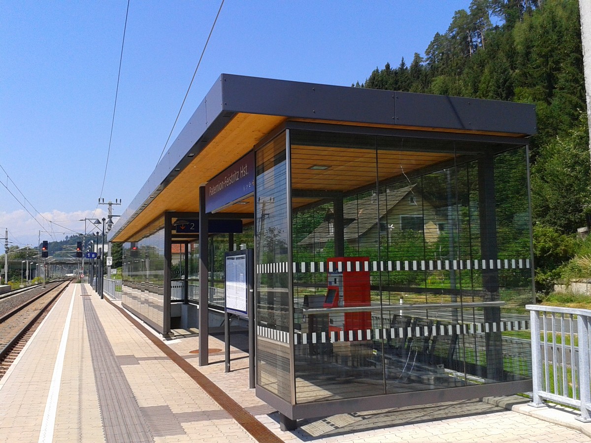 Statt Fahrkartenschalter und Warteraum nurmehr Automat und Unterstand, so sieht es nach der Inbetriebnahme der neuen Haltestelle Paternion-Feistritz aus. Wie zu sehen ist sind hier nur drei Sitze pro Bahnsteig im Trockenen. Ob diese Haltestelle eine Verbesserung für die Kunden darstellt bleibt zu bezweifeln. Der alte Bahnhof ist derzeit nurmehr für die Holzverladung in Betrieb. Der Warteraum wurde schon einige Zeit vor der Inbetriebnahme der Haltestelle zugemauert und als Provisorium vorübergehend ein winziger Unterstand aus Pressholzplatten, mit einem Automaten darunter, gebaut.
