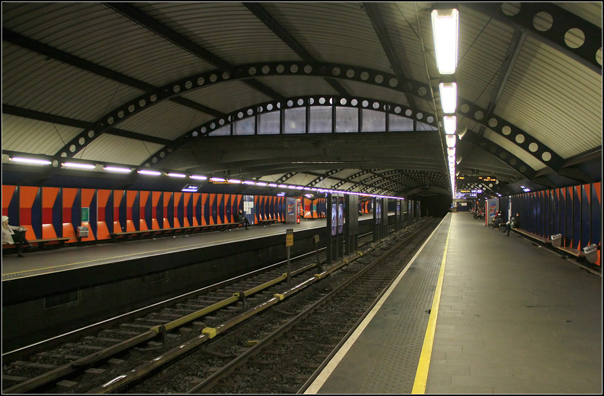Stortinget, der Mittelpunkt des Osloer U-Bahnnetzes -

Nicht immer war das Osloer U-Bahnnetz einheitlich. Früher gab es zwei nicht kompatible Systeme im West und Osten der Stadt, die auch im Zentrum an unterschiedlichen, weit auseinander liegenden Stationen endeten. Das westliche eher eine Vorortbahn mit Oberleitungen und nicht kreuzungsfreien Strecken, einschließlich der Holmenkollenbahn endete seit 1928 unterirdisch in der Station Nationaltheatret. Das östliche Netz, eine echte U-Bahn mit Stromschienen hatte ihren Ausgangspunkt auf der anderen Seite des Zentrums am Jernbanetorget, dem Osloer Hauptbahnhof. Diese 'echte' U-Bahn wurde 1966 eröffnet nachdem vorher schon einzelne Strecken im Straßenbahnvorlauf betrieben wurde.

Es gab dann immer Pläne, die beiden Teilnetze zu verbinden, ohne dass an einen durchgehenden Betrieb gedacht wurde. 1977 wurde das Ostnetz (U-Bahn) nach Westen verlängert bis zur Station Sentrum (heute Stortinget). Dieser wurde 1983 aufgrund von Baumängeln wieder geschlossen und 1987 wiedereröffnet. Zu diesem Zeitpunkt wurden auch die Westlinien (Oberleitungsbetrieb) bis hier her verlängert und endeten in der im Bild gezeigten Bahnsteighalle. Die U-Bahn von Osten her wendete in einer Schleife um diese Bahnsteighalle herum, mit zwei getrennten Bahnsteige für ankommende und abfahrende Züge. Diese Bahnsteige sind direkt über Fußgängertunnels mit den mittleren beiden Gleisen der Westlinien verbunden. Quasi eine viergleisige Station mit zwei Mittelbahnsteigen zum direkten umstiegen in gleicher Fahrtrichtung. 

Ab 1993 wurde ein erster durchgehender Betrieb zwischen den Ost- und Westlinien eingerichtet, der nach und nach auf alle Linien übertragen wurde. Es gab dann Fahrzeuge, die sowohl für Oberleitungs- als auch Stromschienenbetrieb geeignet waren.
Inzwischen sind aber die Westlinien ebenfalls komplett auf Stromschienenbetrieb umgebaut und bis auf die Holmenkollenbahn auf kreuzungsfrei ausgebaut.

Der durchgehende Betrieb findet auf den beiden im Bild gezeigten Gleisen statt, während die Wendeschleife für Verstärkerzüge aus/in Richtung Osten genutzt wird.

Oben schneidet die Fußgängerebene mit einer Art Brücke in die gewölbte Decke der Bahnsteighalle ein.

30.12.2013 (M)