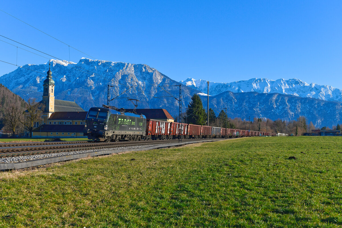 Straff durchgetaktet erwies sich am 25. Februar 2024 der Einsatzplan der für TXLogistik fahrenden 193 212. Kurz vorher erst mit einem KLV-Zug aus Norddeutschland  in Kufstein angekommen, wurde gleich wieder ein Ganzzug, bestehend aus offenen Waggons der Gattung Eaos, in Richtung Deutschland bespannt. Am Kloster Reisach konnte ich den zum Aufnahmezeitpunkt erst kürzlich neu gestalteten Werbe-Vectron zum zweiten Mal an diesem Tage aufnehmen. Während im Inntal schon frühlingshafte Temperaturen herrschten, zeigten sich der Wilde und der Zahme Kaiser im Hintergrund frisch verschneit. 