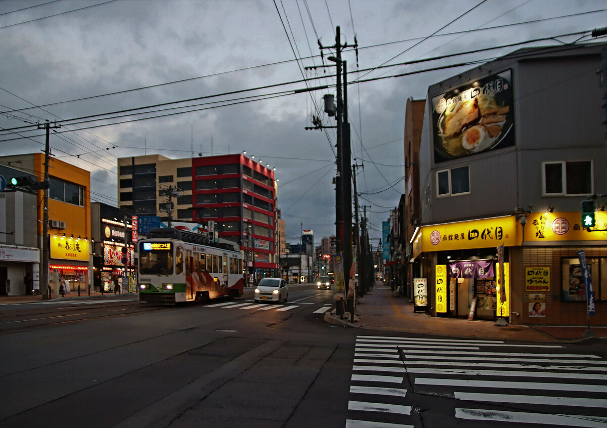 Strassenbahn Hakodate im Süden der Insel Hokkaidô, Japan: Langsam wird es Nacht und die kleinen Restaurants leuchten auf, während Wagen 3002 sich dem Bahnhof nähert. 29.Oktober 2022. Wagen 3002  entstammt einer Serie von 4 Wagen mit Nutzbremse, 3001-3004, von denen je 1 Wagen 1993, 94, 95 und 96 gebaut wurde.  