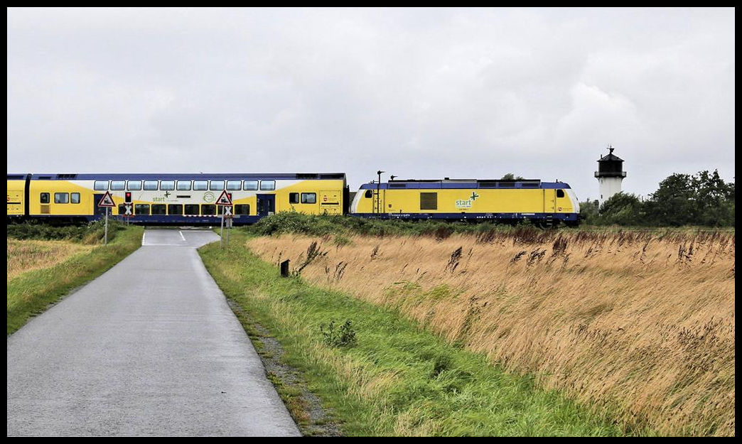 Südlich von Cuxhaven passiert hier an 17.08.2021 um 12.19 Uhr der Start Zug mit Lok 246006-1 nach Hamburg den Bahnübergang nahe des Leuchtturm Dicke Berta.