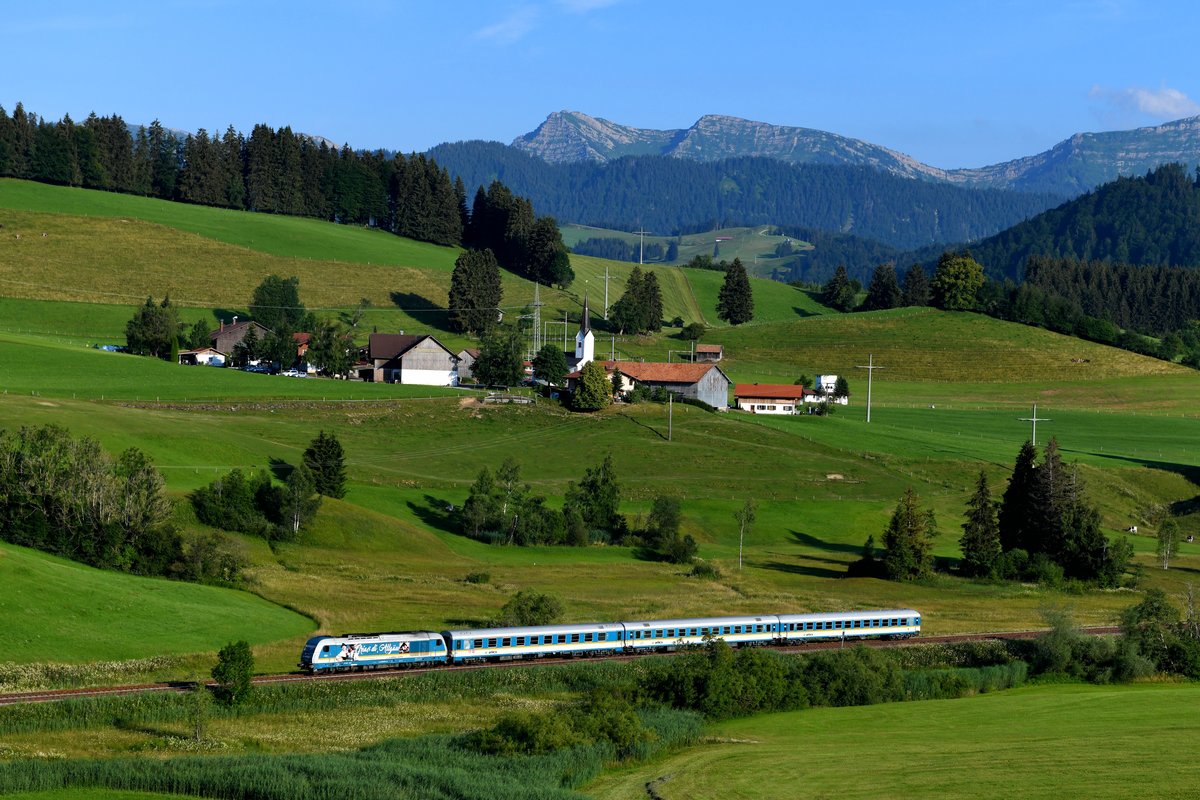 Tagesabschluss für den 01. Juli 2018 bildete der ALX 84142 nach Lindau. Diesen konnte ich vor der kleinen Ortschaft Zell mit dem Rindalphorn und dem Gelchenwanger Kopf im Hintergrund fotografieren. Es führte die 223 072, die zum Aufnahmezeitpunkt eine Seitenwerbung trug, welche auf das „Gautrachtenfest D’Wageggler“ hinweisen sollte. Mit einem Trachtenpaar und der Aufschrift  Griaß di, Allgäu!  möchte man die Verbundenheit zur Region unterstreichen. 