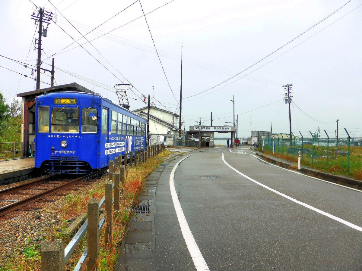 Takaoka Manyô-Linie: Die Linie endet hier nach 13 km Fahrt in Koshinokata abrupt, da die in den 1960er Jahren gebauten neuen Hafenanlagen die Landverbindung Richtung Toyama gebrochen haben. Im Hintergrund ist die Anlegestelle der Fähre erkennbar. Im Bild der alte Wagen 7076, 25.Oktober 2013. 