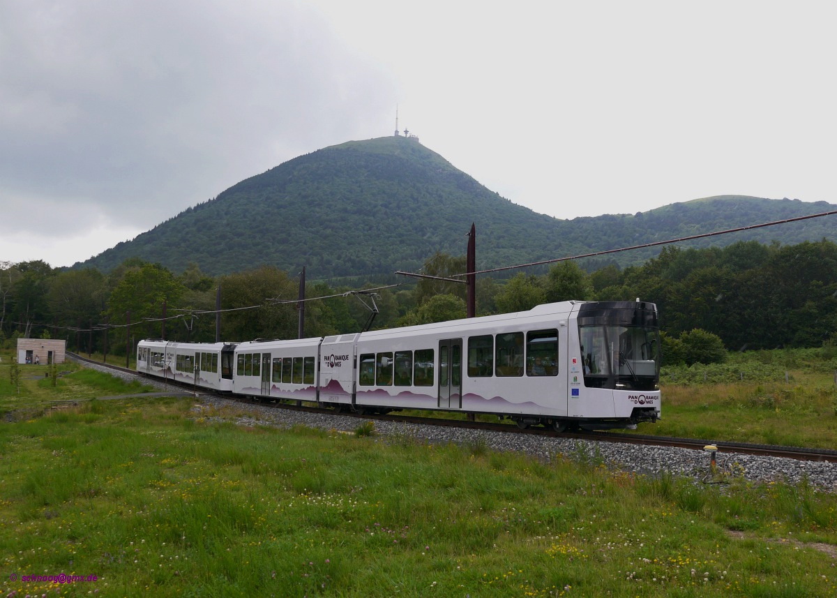 Talfahrt des Beh 2/6 1 und 2 vor dem Hintergrund des Gipfels des Puy de Dôme.
 
Seit Mai 2012 fährt die neuerbaute Zahnradbahn Panoramique des Dômes zum Gipfel des Puy de Dôme.
Die 5,3 Kilometer lange Strecke führt von der Talstation (Maison de Site) zur Gipfelstation (Gare de Sommet) auf 1406 Meter Höhe. Die meterspurige Strecke hat Y-Stahlschwellen und eine Zahnstange nach System Strub. Die vier Triebwagen Beh 2/6 (Typ GTW) wurden von der Firma Stadler in der Schweiz gebaut.

Der 1465 Meter hohe Puy de Dôme ist ein Vulkangipfel, der die zu seinem Fuße liegende Großstadt Clermont-Ferrand um gut 1000 Meter überragt.


2014-07-26 Puy de Dôme
