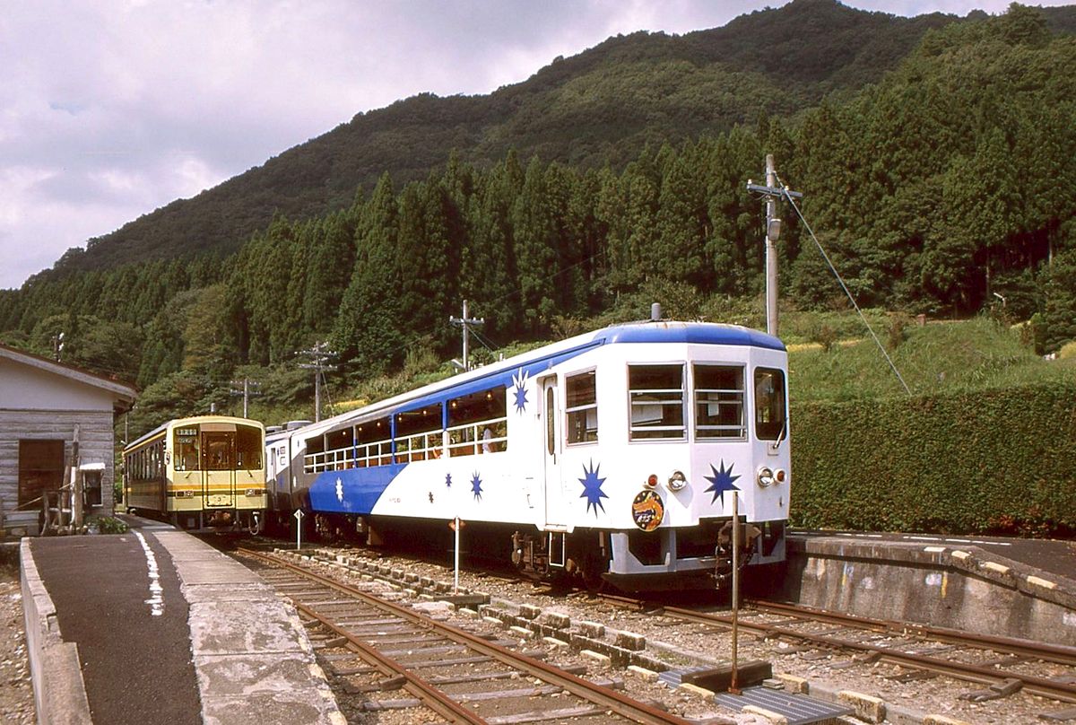 Touristikzug auf der kaum befahrenen Kisuki-Linie: Der Zug ist über eine Spitzkehre hoch oben im Wald Steuerwagen voran nach Izumo Sakane heruntergekommen. Jetzt macht er wieder eine Spitzkehre und wird dann Lok voran weiter in die Ebene hinunterfahren. Links wartet einer der 3 Regelzüge des Tages (Triebwagen KIHA 120-3)auf die Weiterfahrt bergauf. 21.September 2002. 