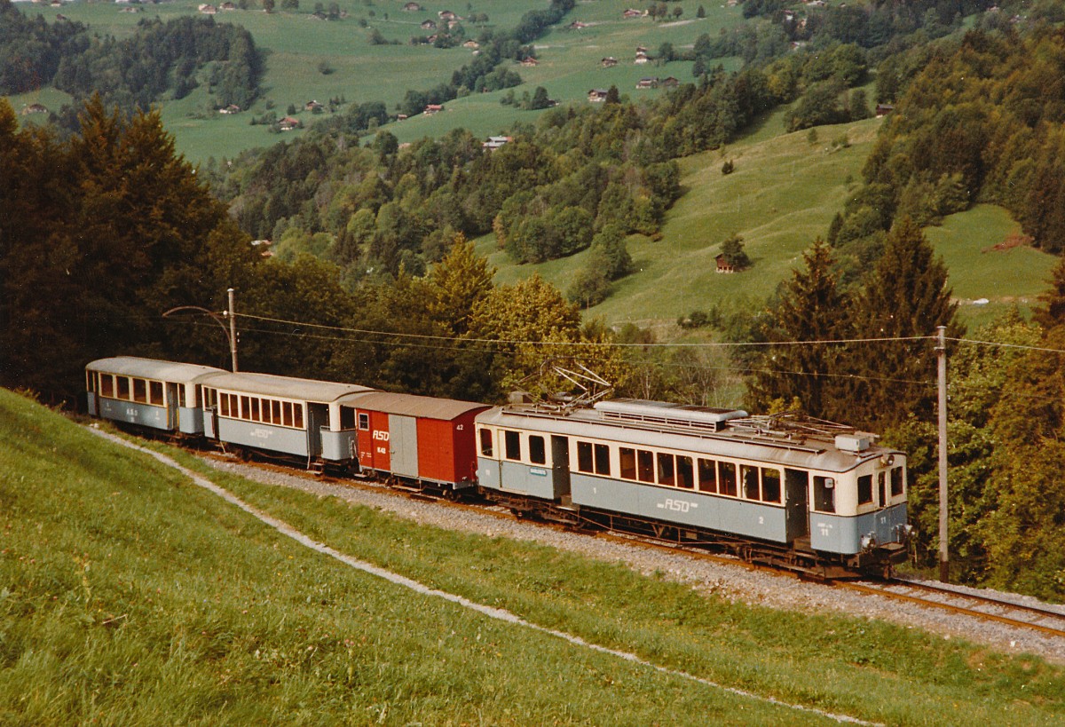 TPC/ASD: 100  Jahre ASD. Ueber das kommende Wochenende feiert die ASD ihren 100. Geburtstag. Zu diesem Anlass wird der restaurierte BCFe 4/4 1 im grau-weissen Anstrich mit Nostalgiezügen im Einsatz stehen. Gemischter Zug mit dem ABFe 4/4 11 oberhalb Les Planches im Oktober 1982.
Foto: Walter Ruetsch   