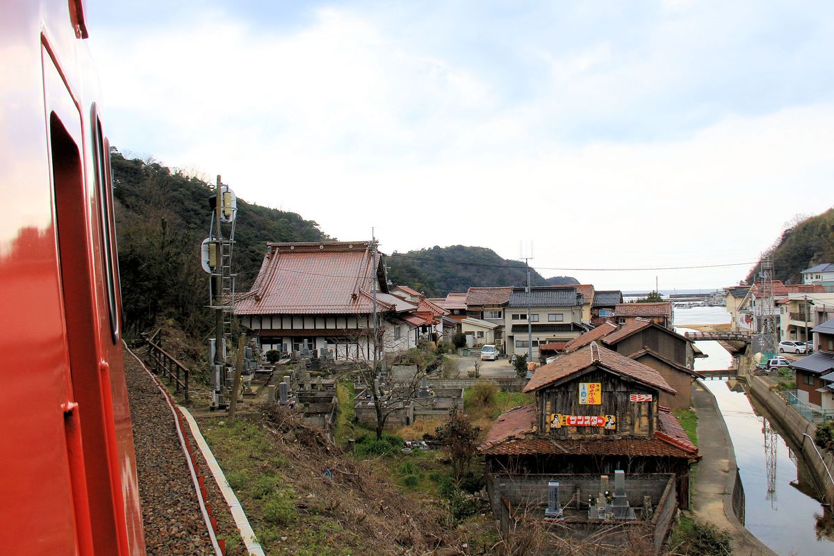 Triebwagen KIHA 47 2018 von JR Westjapan fährt in Yunotsu ein. Das Fischerdörfchen, das auch für seine heissen Quellen bekannt ist, wirkt verlassen, uralte Häuser, ein Tempel und im Vordergrund einige Gräber, hinten das Japanische Meer. 26.März 2017.  