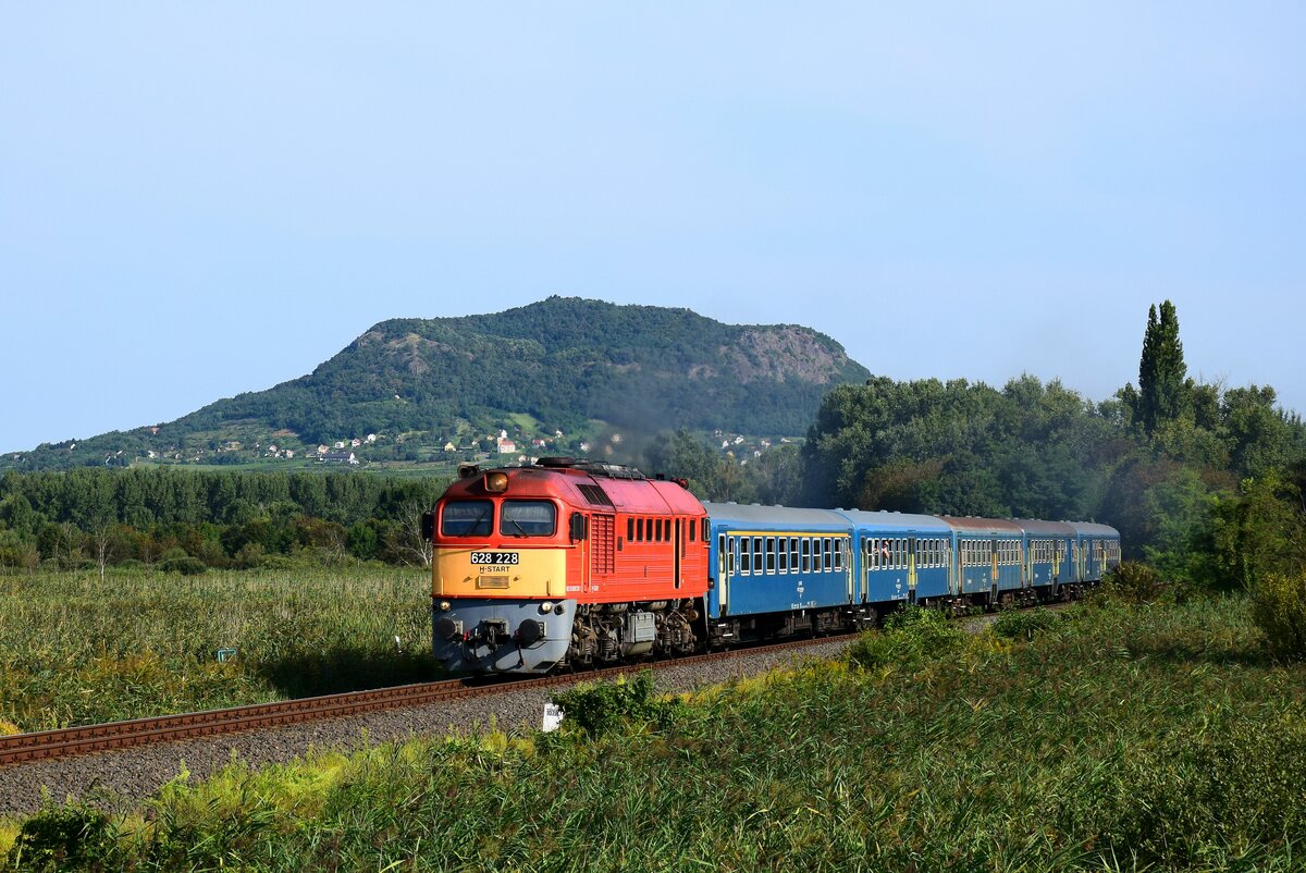 Typische Balaton-Landschaft. Die 628 228 (ex. M62 228) ist mit dem Zug 19787 (Tapolca-Balatonfüred) unterwegs kurz nach Bf. Badacsonytördemic-Szigliget mit dem Szent György Berg im Hintergrund.
27.08.2023.