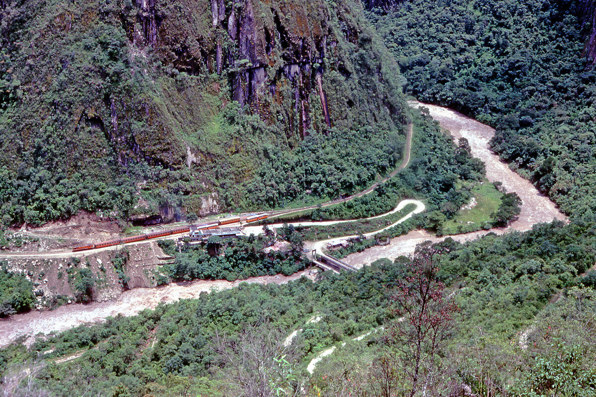 �bersichtsbild bei meinem ersten Besuch im Januar 1980, gemacht von der alten Inkast�tte Machu Picchu aus (ca 2500 m�M): Der Zug steht im alten Bahnhof Puente Ruinas (1859 m�M). Die Serpentinenstrasse f�hrte zuerst �ber den Fluss Urubamba und dann dem Berg entlang nach oben. Parellel dazu gab es (und gibt es immer noch) einen Fusspfad f�r die sportlichen Touristen. Rechts hinter der Kurve befindet sich das Dorf Aguas Calientes (ca 2000 m�M), dort befand sich im Jahr 2015 der Personenbahnhof. Bei unserem Besuch am 28.1.1980 war die Serpentinenstrasse zum Machu Picchu hinauf ein paar Tage zuvor versch�ttet worden, das hiess, die Inkastadt war nur zu Fuss erreichbar. Dies kam uns als fitte Burschen entgegen, wir waren als erste oben und konnten so die ganze Ruinenstadt ohne Touristen erkunden und fotografieren. Gl�ck hatten wir dennoch, denn ab dem folgenden Tag verkehrte der Touristenzug nicht mehr, weil ja die Busse ab dem Bahnhof hinauf zu Ruinenstadt Machu Picchu nicht mehr verkehren konnten. Machu Picchu, 28.1.1980