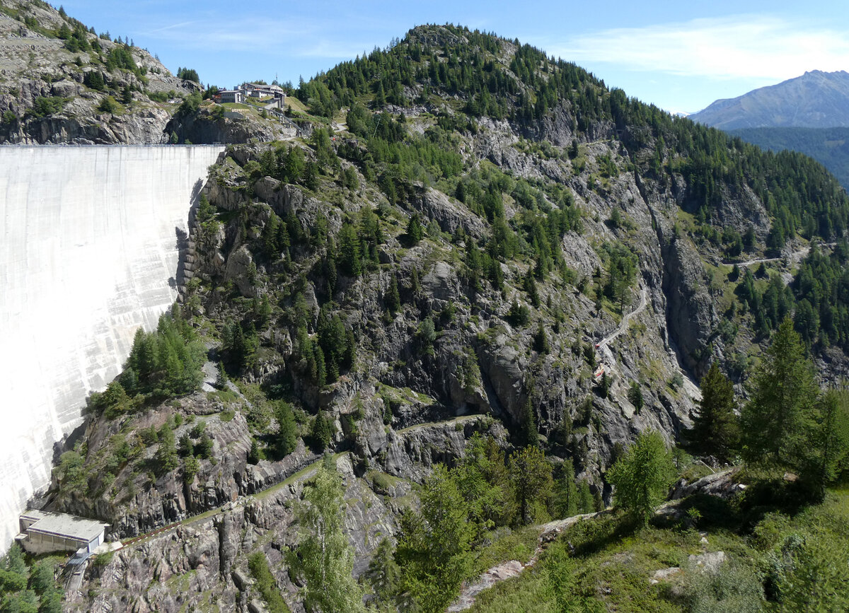 'Übersichtsfoto' vom Trassee des Panoramic Train zum Lac d'Emosson, dessen Staumauer links teilweise sichtbar ist. Rechts hinten erscheint der Panoramazug jeweils von Les Montuires und fährt durch einen Tunnel hinter dem ersten Felsen. Vor dem zweiten Tunnel steht der Panoramic Train im Endbahnhof Pied du Barrage, dort steigen die Fahrgäste auf eine Art Schräglift um. Diese führt sie zu den Gebäuden oberhalb der Staumauer (Bergstation, Restaurant etc.). Dort befindet sich auch eine Busstation, aber die Anreise auf der Strasse von Finhaut ist natürlich bei weitem nicht so spektakulär wie jene mittels Standseilbahn, Panoramic Train und minifunic. Links der Station Pied du Barrage geht das Trassee durch zwei Tunnels weiter in Richtung Staumauer, allerdings ohne Gleis. Links unten befinden sich ein paar Meter Gleis, dieses führt um eine Kurve in ein weisses Gebäude am Fusse der Staumauer. Ob es sich dabei um ein altes Depot handelt, ist mir nicht bekannt. Das Foto wurde von der gebogenen Staumauer aus gemacht. Lac d'Emosson, 25.8.2025