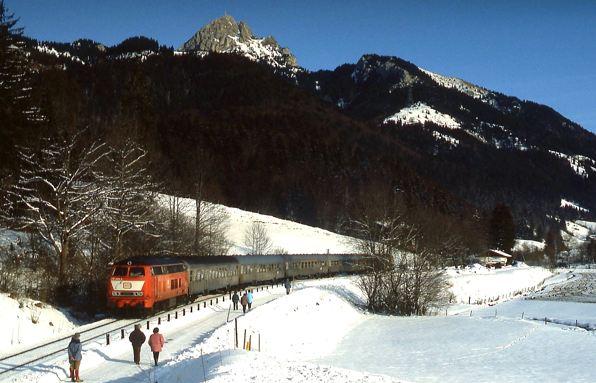 Unter den Augen der Spaziergänger, die das herrliche Winterwetter zu einem Ausflug nutzen, verlässt eine 218 Ende Dezember 1991 mit einem Zug von Bayrischzell nach München den Haltepunkt Geitau.