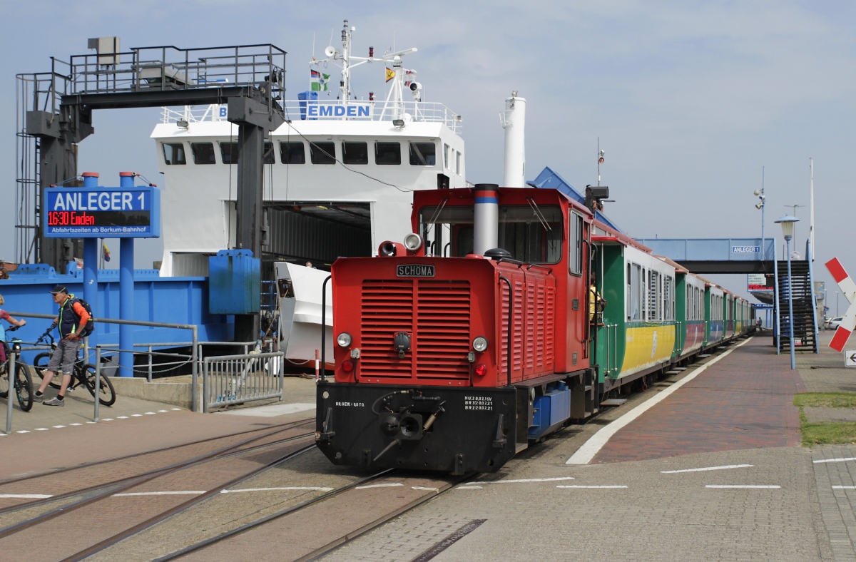 Vor einem Urlauberzug wartet die Lok  Berlin  der Borkumer Kleinbahn am 26.06.2021 auf die Abfahrt in Richtung Borkum Kleinbahnhof, im Hintergrund die Fähre  Ostfriesland 