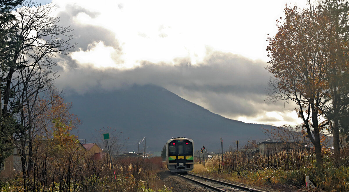 Vor kaum einer Minute zeigte sich der Vulkan Yôteizan im mittäglichen Sonnenschein. Doch als Triebwagen H100-7 sich dem Einfahrtsignal von Kutchan nähert, ziehen wieder schwere Regenwolken über den Berg. 29.Oktober 2022 