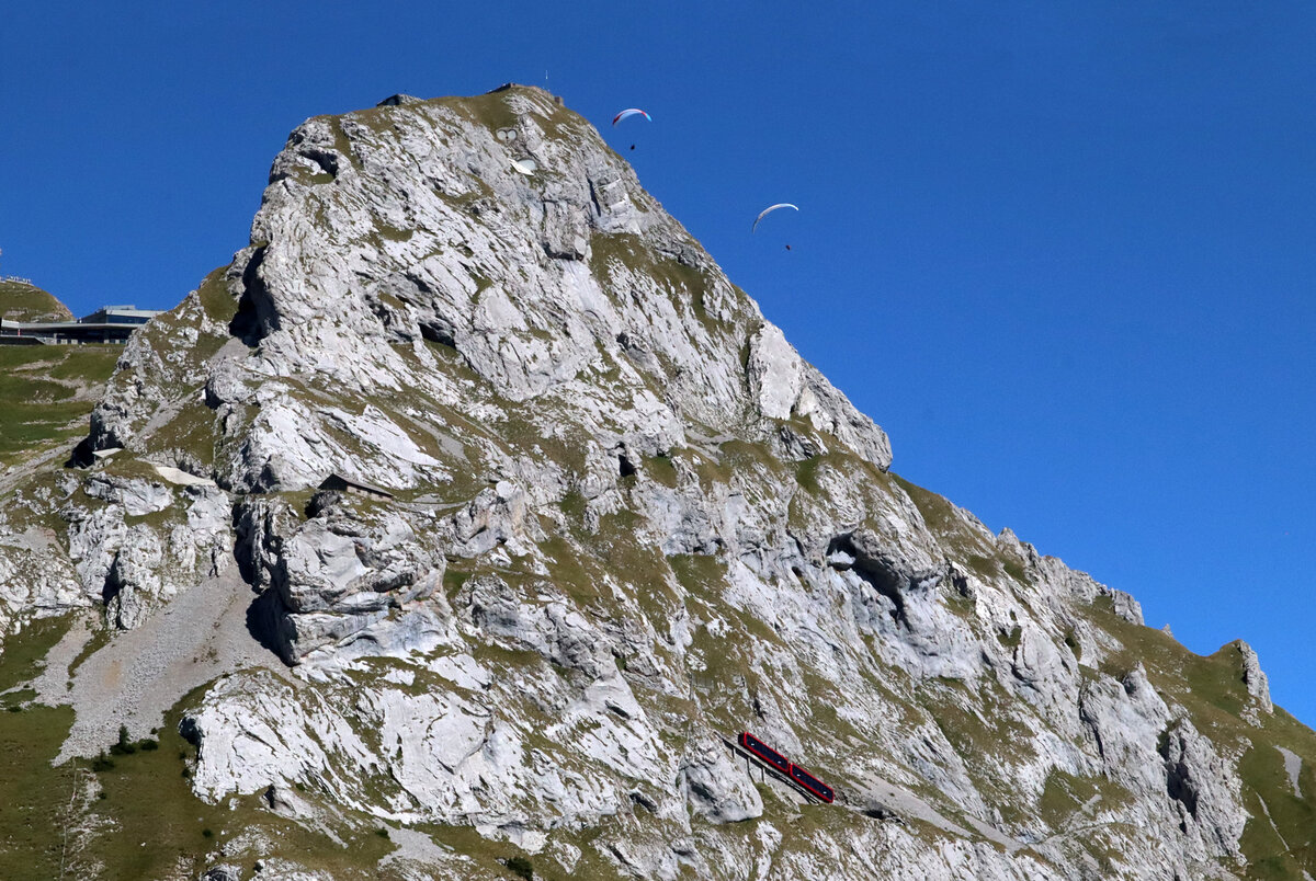 W�hrend der Doppeltriebwagen in der Eselswand bergw�rts f�hrt, sind zwei Gleitschirmflieger in der Luft unterwegs. Das Foto wurde vom gegen�berliegenden Hang aus gemacht. Pilatus Kulm, 19.8.2026