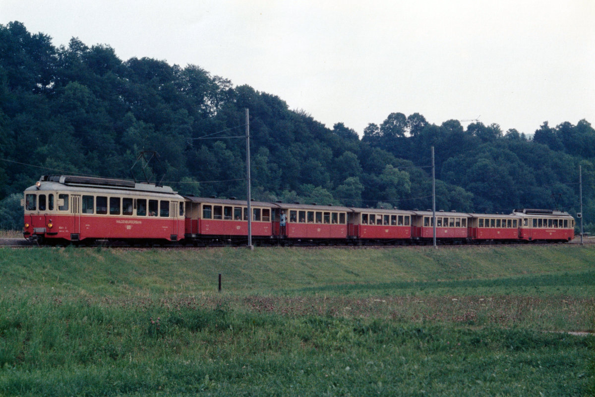 WALDENBURGER BAHN/WB.
GROSSER ZUG AUF KLEINER BAHN.
Mit etwas Fotografenglück erwischte man auch ab und zu einen langen Zug auf der  KLEINEN BAHN .
Regionalzug bestehend aus sieben Elementen im Juni 1980 zwischen Niederdorf und Hölstein auf der Fahrt von Waldenburg nach Liestal. Besondere Beachtung gilt den BDe 4/4 Motorwagen an beiden Enden dieses Zuges sowie den Wagen mit offenen Blattformen.
Foto: Walter Ruetsch