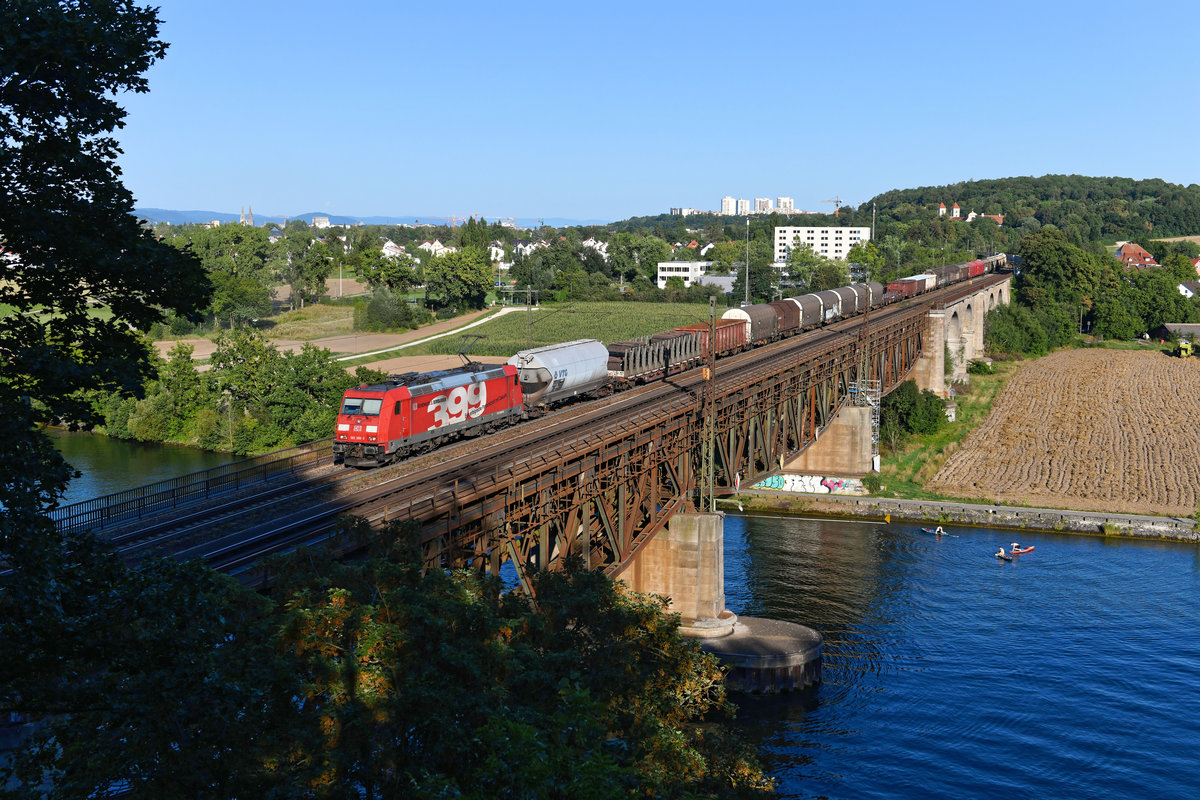 Wenige Augenblicke, bevor die Fotostelle im Schatten lag, überquerte am 21. August 2020 noch die sogenannte Plattlinger Übergabe die Mariaorter Eisenbahnbrücke. Als Zuglok war an diesem Tag die 185 399 eingeteilt, die mit ihrer seitlichen Beschriftung auf die enge geschäftliche Beziehung des Herstellers Bombardier zur Güterzugsparte der Deutschen Bahn hinweist. 