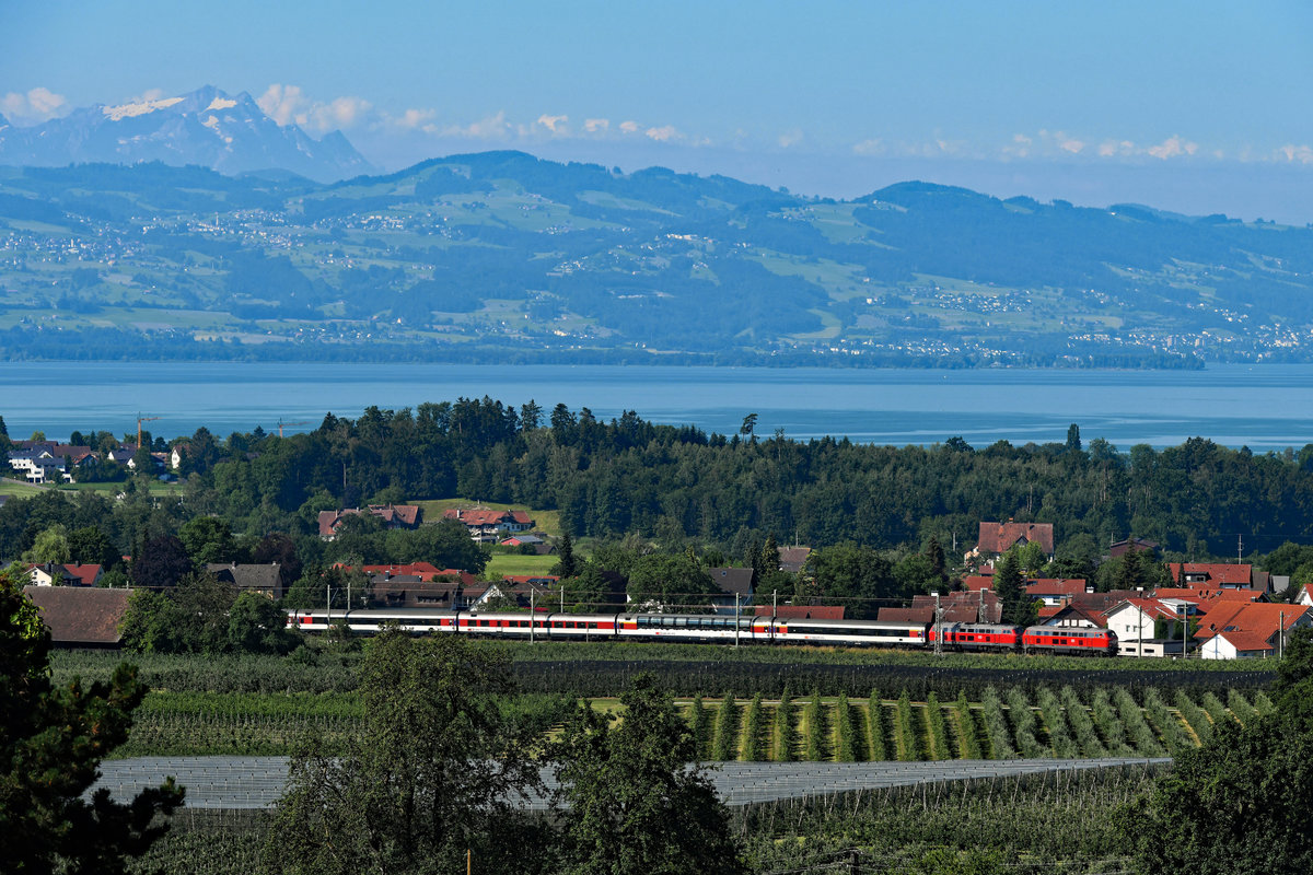  Wer zu spät kommt, den bestraft das Leben - oder man muss mit Masten leben.  Inzwischen ist die Elektrifizierung des letzten Abschnitts der KBS 970 schon weit voran geschritten und die Oberleitung steht. Gerade in der Gegend um Bodolz, wo sich die Allgäubahn serpentinenartig durch die Obstplantagen windet, sind viele Motive leider unbrauchbar geworden. Ganz zu spät war ich zu Sommerbeginn diesen Jahres aber doch nicht dran, zumindest stand die Lärmschutzwand noch nicht. Von Taubenberg aus hat man diesen schönen Blick auf den Ort Bodolz, den Bodensee und das Ufer auf Schweizer Seite bei Rorschach sowie den Appenzeller Ausserrhoden. Über allem thront der 2.501 Meter hohe Säntis. Am 25. Juni 2020 konnte ich dort die 218 452 und 403 aufnehmen, die an diesem Tage den EC 191 von Lindau nach München HBF brachten. 