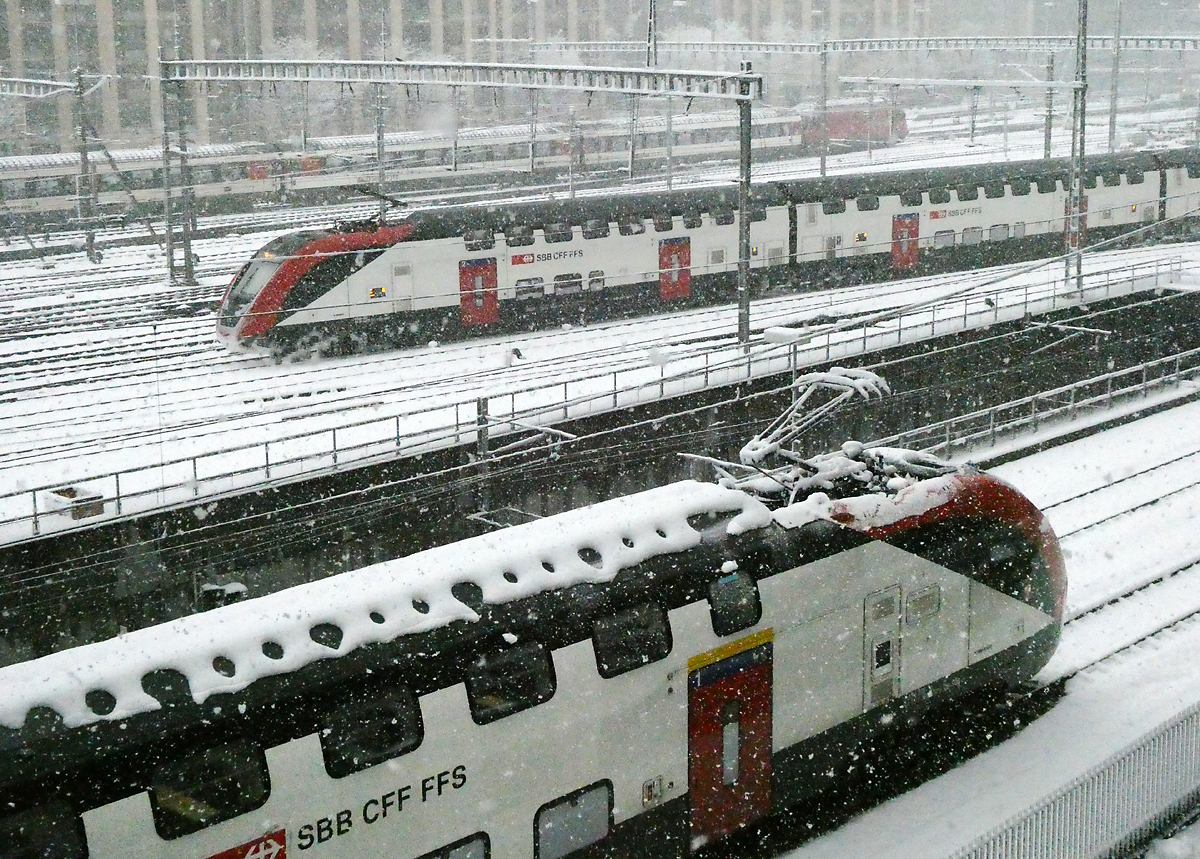 Zürich HB: Blick vom Negrellisteg auf das Gleisfeld und das Schneetreiben. Gemäss dem Schnee auf dem Pantografen auf dem FV-Dosto im Vordergrund scheint dieser Zug schon einige Zeit dem Schneefall ausgesetzt zu sein. Im Hintergrund fährt ein Zug derselben Bauart in den Bahnhof ein. Im Einschnitt in der Bildmitte führen zwei Gleise in den unterirdischen Bahnhofteil 'Löwenstrasse'. Zürich HB, 2.12.2023