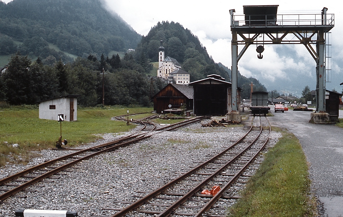 Zwischen 1927 und 1961 betrieben die Vorarlberger Illwerke eine 760 mm-Schmalspurbahn, die vom Bahnhof Tschagguns an der Montafonerbahn nach Partenen führte. Sie diente dem Materialtransport zu den beiden Kraftwerken in Vermunt und Obervermunt und dem Silvrettastausee. Der planmäßige Betrieb, auch Personenverkehr, endete 1953. Nach der Einstellung wurden die Gleise bis auf ein etwa 500 m langes Reststück, das bis 2001 zur gelegentlichen Materialanlieferung für die Illwerke diente, abgebaut. Teilweise wurde der Oberbau für den Ausbau der Stainzerbahn verwendet. Bis 2011 wurde in dem in der Bildmitte sichtbaren Lokschuppen das Motorboot  Silvretta , das im Sommer auf dem Silvretta-Stausee verkehrte, im Winter abgestellt. 2013 wurden die noch vorhandenen Anlagen abgebaut. Die Aufnahme entstand im August 1982 in Tschagguns.