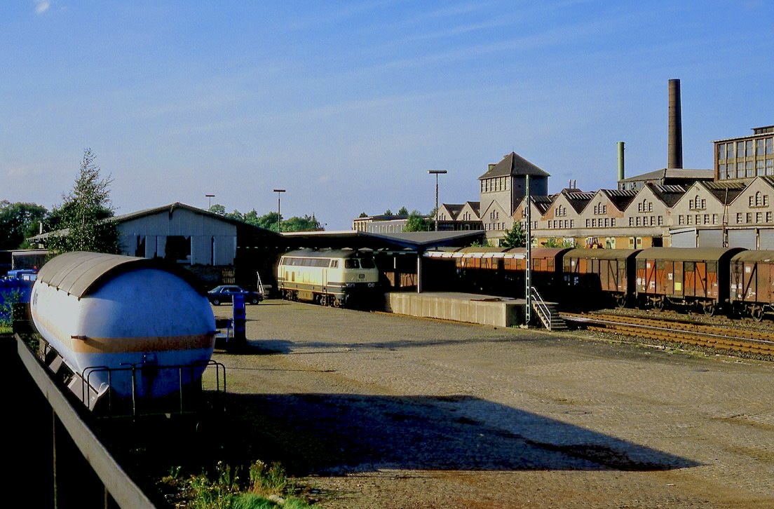 215 021 an der Güterrampe des Bf.Velbert, 02.09.1985. Nachdem die Schnellstraße Velbert - Wuppertal eröffnet worden war, wurde die Stückgutverladung nach Wuppertal Langerfeld verlegt. Die beiden Zugpaare sind entfallen, der Bahnhof wurde zurückgebaut. Anstelle der Gießerei im Hintergrund gibt es nun einen riesigen Baumarkt, der auch große Teile des Bahngeländes nutzt.