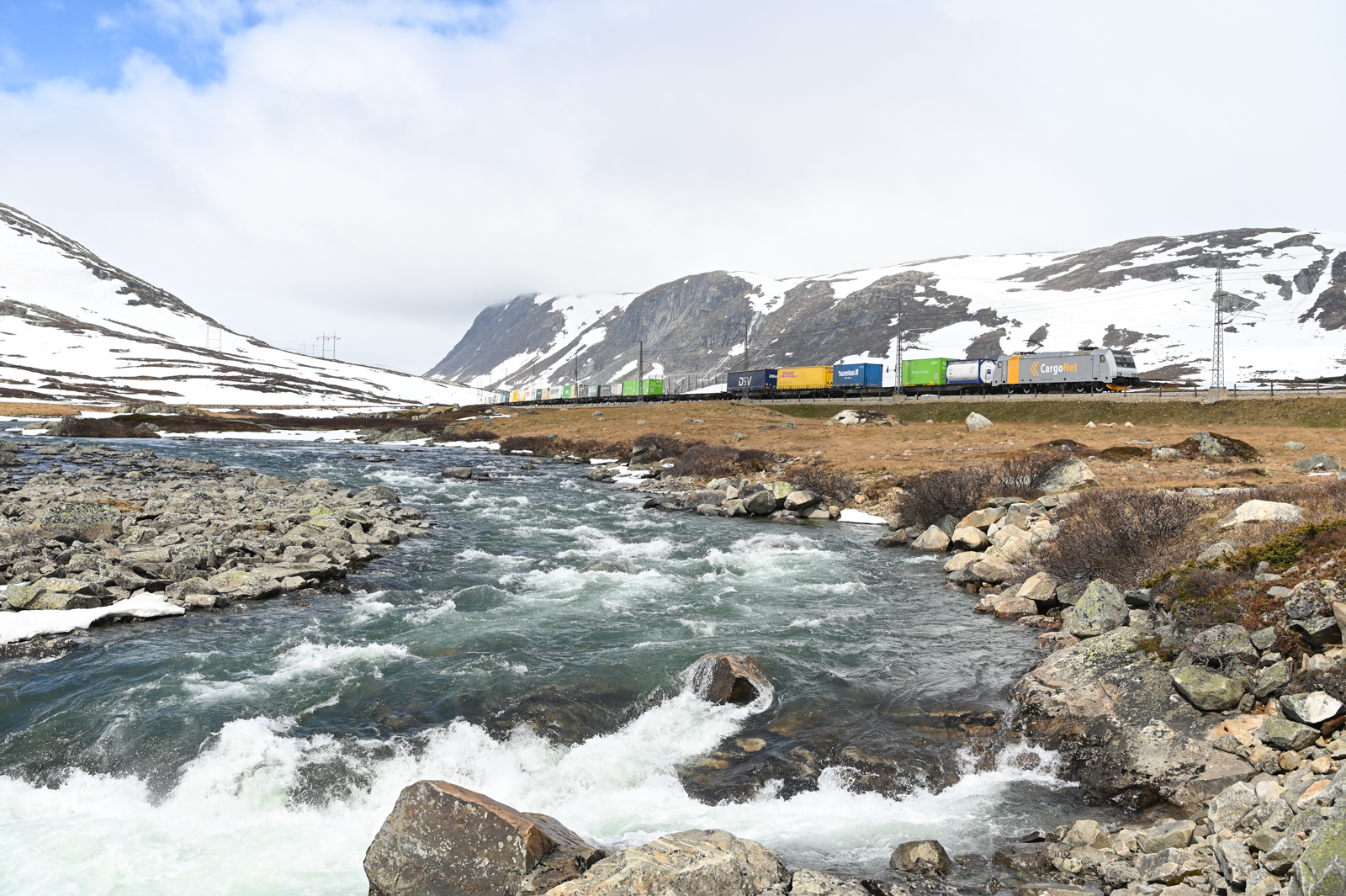 Am Vormittag des 03.06.2022 hängen noch ein paar Wolken an den Bergen der Hardangervidda, einer Hochebene in Mittelnorwegen. Entlang der einsamen Bergwelt und vorbei an Wasserfällen fährt der Güterzug 5506 seinem Ziel Oslo entgegen. Hier zwischen Finse und Haugastøl.