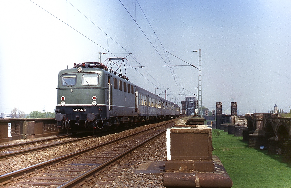 Anfang der 1980er Jahre fährt 141 156-0 mit einem Nahverkehrszug über die Neusser Rheinvorlandbrücken der Rheinbrücke zwischen Düsseldorf und Neuss. Rechts sind die Reste der nach dem 2. Weltkrieg nicht wieder aufgebauten südlichen Eisenbahnbrücke zu sehen. Dort steht heute die neue viergleisige Brücke.