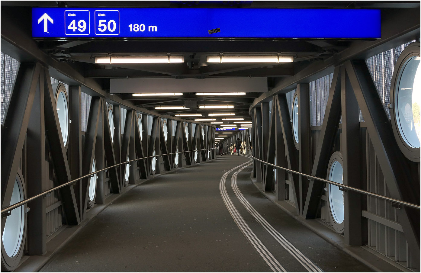 Der Bahnhof Bern -

Die Fahrgastbrücke zu den 180 Meter entfernten Gleisen 49 und 50 am verlängerten Bahnsteig der Gleise 9 und 10. Der Schwung am Beginn des Stege erklärt sich aus Postion über den Gleisen 8 und 9 im Anschluss an die 'Welle von Bern.' So musste nicht in die Bahnsteigdächer eingegriffen werden. Im weiteren Verlauf folgt die Brücke dem Bahnsteig, der wiederum eine Krümmung aufweist.

07.03.2025 (M)