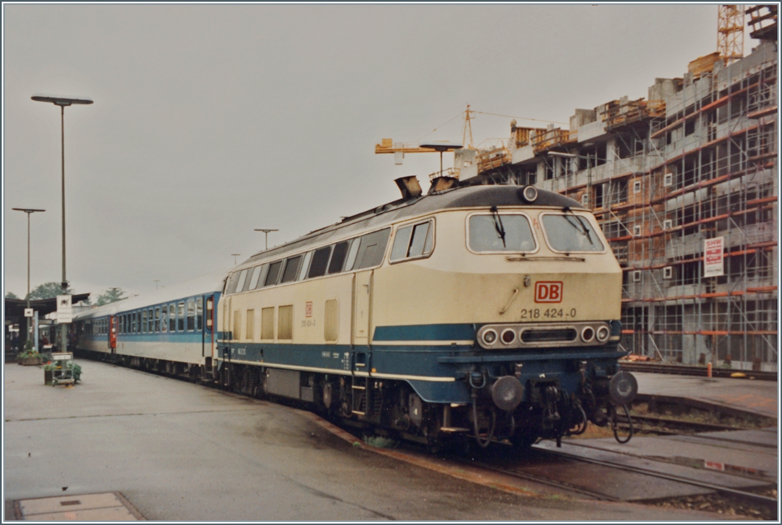Die DB 218 424-0 hat in Friedrichshafen auf Gleis 3 den IR 360 übernommen, Richtungswechsel: an 13:26/ab 13:33. Der Zug der IR Linie 26 ist von Bregenz (ab 12:49) nach Saarbrücken (an 18:10) unterwegs. Der Zug ist als klassische IR Komposition gereiht: Bimd 255, Bim 256, Bim 257, Bim 258, Bim 259, ARkimbz 260 und dem Ami 261. Von Bregenz nach Lindau HBF verkehrte der Zug als D 360. 
Rechts im Bild entstehen Wohngebäude die bis heute den Hintergrund des Bahnhofs von Friederichshahen bestimmen.

Analogbild vom 30. Mai 1995