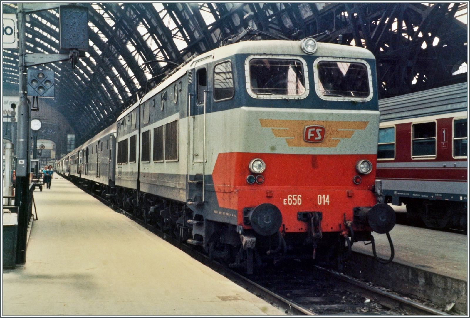 Die FS E 656 014  Caimano  ( Kaiman  Alligator/Krokodil) wartet in Milano Centrale mit einen typischen Reisezug aus grauen Wagen auf die Abfahrt, rechts im Bild ist ein FS Wagen in einer neuen Farbvariante zu erkennen.  

Analogbild vom Juni 1985