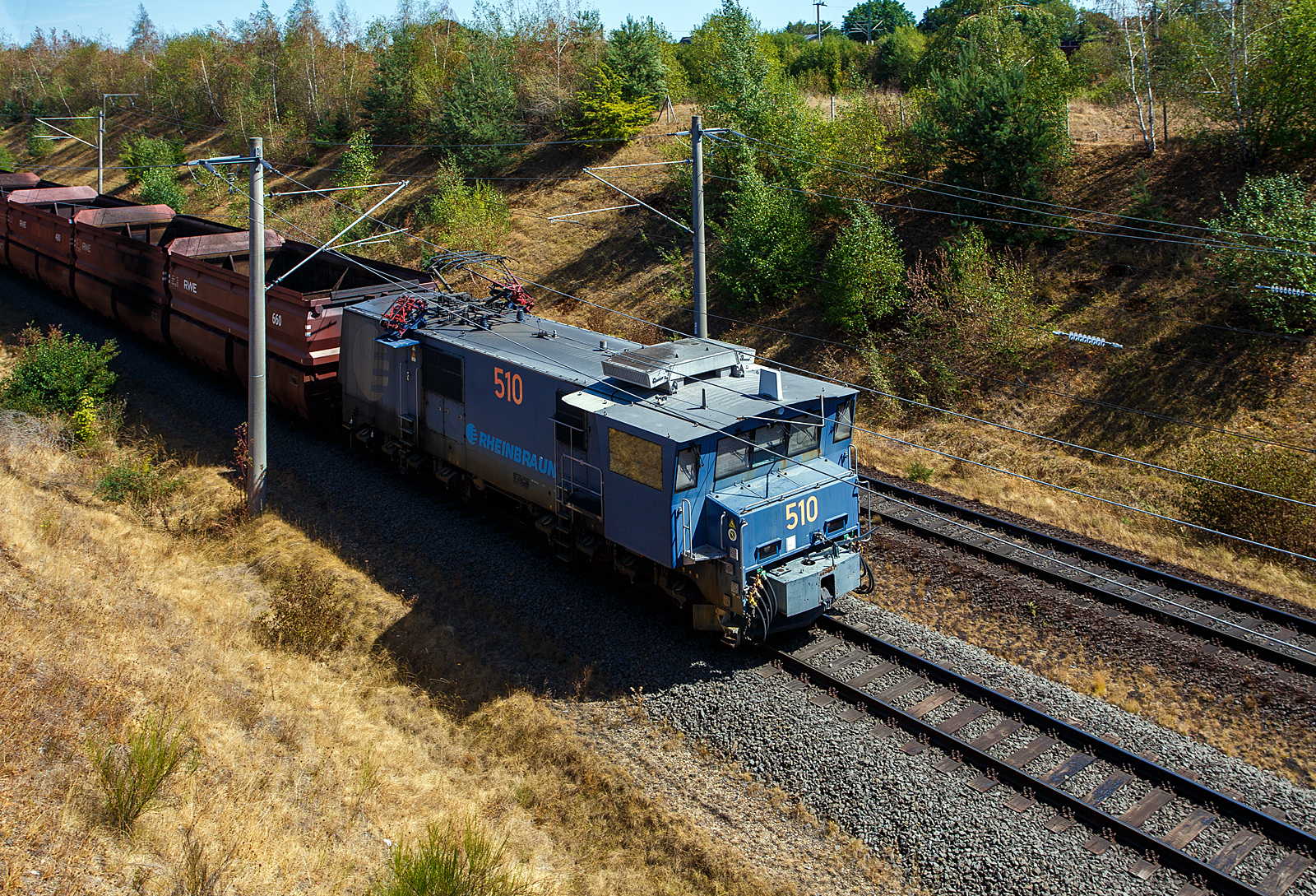 Die Tagebaulokomotive Adtranz - EL 2000 – 510 der RWE Power AG (ex Rheinbraun) kommt am 12 August 2026 bei Kerpen-Buir auf der Hambachbahn mit einen Braunkohle Selbstentladewagen Leerzug, von den Kraftwerken zum Tagebau Hambach.

Die RWE Power AG setzt zwei verschiedene Elektrolokomotiven ein - die zwischen 1954 und 1966 in gro�er St�ckzahl durch verschiedene Hersteller (Henschel, Krauss-Maffei und Krupp) gelieferte Type EL 1 (aktuell 21 Fahrzeuge im Einsatzbestand), sowie die 1999 - 2000 vom Kasseler ADtranz-Werk in einer St�ckzahl von zehn Maschinen gelieferte EL 2000. Die RWE Lok 510 wurde 2000 ADtranz (ABB Daimler Benz Transportation Adtranz) unter der Fabriknummer 33327 gebaut und an die Rheinbraun AG geliefert. Bei RWE Power hat sie zudem die Equip-Nr. 40043.

Adtranz EL 2000 sind die etwas st�rkeren Nachfolger der EL 1 Lok, da diese in die Jahre gekommen waren (Bj1954 bis 1965), musste Ersatz beschafft werden. Die elektrischen Tagebaulokomotiven des Typs Adtranz EL 2000 sind typischerweise (wie auch die EL 1) mit einer robusten Mittelpufferkupplung mit breiter Aufschlagfl�che ausger�stet. Diese Spezialkupplung ist notwendig, um den harten Belastungen beim Verschieben schwerer Abraum- und Kohlez�ge im rheinischen Braunkohlerevier standzuhalten. Markant ist neben der Kupplung das seitlich �berkragende F�hrerhaus f�r eine bessere Streckensicht bei geschobenen Z�gen. Auch die vielen Sandbeh�lter, je Seite sind es 8 St�ck sind sehr auff�llig. Neben den zwei Einholmstromabnehmer (im Tandem), besitzen die Tagebauloks seitliche Stromabnehmer, diese werden an der Verladestation (Kohlebunker) ben�tigt, damit die Waggons ohne st�renden Fahrdraht beladen werden k�nnen.

Technik
Die Lokomotiven besitzen einen F�hrerstand auf einer Seite mit einem kleinen Vorbau davor. Der F�hrerstand ist �ber 1,3 m breiter als der Fahrzeugkasten. Dadurch kann die Lokomotiven auch im Betrieb mit dem Maschinenraum voran betrieben werden. Die der Fahrleitung entnommene Energie (6 kV 50 Hz) wird auf dem Fahrzeug in die f�r die Fahrmotoren (Typ: ABB 6 FRA 7072) geeigneten umgewandelt. Dies geschieht mittels Trafo, 4 QS-Steller, Spannungszwischenkreis und Wechselrichter. Die Fahrmotoren sind �hnlich denen der bekannten Doppel-Elektrolok „IORE“ der schwedischen Erzbahn LKAB Malmtrafik AB. Die Antriebsleistung von den Fahrmotoren wird jeweils �ber Tatzlager mit einer �bersetzung 6,266 : 1 auf die Antriebwellen (Radsatzwellen) �bertragen. Daher sind dann auch die gewaltigen Zugkr�fte m�glich. 

TECHNISCHE DATEN der Adtranz EL 2000:
Gebaute Anzahl: 10 St�ck
Nummerierung: 501 bis 510
Spurweite: 1.435 mm (Normalspur)
Achsfolge: Bo'Bo'
L�nge �ber Puffer: 16.400 mm 
Drehzapfenabstand: 8.200 mm
Achsabstand im Drehgestell: 2.700 mm
Treibraddurchmesser: 1.250 mm (neu)
Breite: 4.500 mm
H�he: 4.075 mm (bei eingezogenem Stromabnehmer)
Eigengewicht: 140 t
Achslast: 35 t
H�chstgeschwindigkeit: 70 km/h
Stromsystem: 6 kV 50 Hz Einphasenwechselstrom
Dauerleistung: 2.800 kW
Anfahrzugkraft: 500 kN
Fahrmotoren: 4 St�ck (Typ: ABB 6 FRA 7072) / je 227 A / je 714 kW
Antriebart: �ber Tatzlager mit einer �bersetzung 6,266 : 1
Strom�bertragung: Oberleitung oder Seitenfahrleitung
Bremsen: Sab Wabco Druckluft-Klotzbremse, Federspeicher-Feststellbremse, Elektro-Bremse mit Stromr�ckspeisung in Fahrleitungsnetz. 
Sicherheitseinrichtungen: SIFA, magnetische Zugbeeinflussung (MZ), mech. Fahrwegsicherung (Druckluft-Bruchrohre), Druckluftpfeife und elektrisches L�utwerk.

Nochmals meinen Dank an Herrn Thomas Nie� der RWE Power AG f�r die Zusendung von Technischen Daten der Loks und Wagen. 

Der Eisenbahnbetrieb der RWE Power AG (ex Rheinbraun)
Auf einem 230 km langen Streckennetz, das auf den Hauptabfuhrstrecken f�r eine Achslast von 35 t ausgelegt ist, werden zu Spitzenzeiten die angeschlossenen Kraftwerke t�glich mit 220.000 – 230.000 t Kohle (das entspricht ungef�hr 170 Zugpaaren) versorgt. Allein das Kraftwerk Niederau�em verbraucht bis zu 90.000 t Kohle t�glich.

Der Eisenbahnbetrieb hat 270 Mitarbeiter, davon 180 Lokf�hrer. Bis zur Einstellung des Abraumbetriebes im Jahre 2001 standen f�r den Gesamtverkehr 54 Elektrolokomotiven zur Verf�gung. Aktuell stehen 31 Elektrolokomotiven im Einsatz. 17 Dieselloks sind vorrangig f�r Infrastrukturinstandhaltung im Einsatz. Daneben werden sie f�r �bergabefahrten in das �ffentliche Netz vorgehalten.

Es stehen 333 vierachsige Kohlewagen mit einem Gesamtgewicht ca. 120 t (bis 114 m�) und 140 achtachsige 96 m� Abraumwagen mit einer Tragf�higkeit von 180 t zur Verf�gung.

F�r Bereisungszwecke steht ein MAN-Schienenbus zur Verf�gung, der von der AKN stammt. Er erhielt von Nov. 2008 bis April 2010 in Delitzsch (EuroMaint) eine umfangreiche Aufarbeitung.


Hambachbahn
Die Hambachbahn ist eine Tagebau-Werksbahn der RWE Power (ex Rheinbraun) im Rheinischen Braunkohlerevier. Sie bindet den Tagebau Hambach an die Nord-S�d-Bahn an, �ber die die RWE-Kraftwerke in Niederau�em, Frimmersdorf und Neurath mit Braunkohle versorgt werden.

Die 1983 in Betrieb genommene Hambachbahn wird, wie auch die Nord-S�d-Bahn, elektrisch betrieben, jedoch als Besonderheit abweichend zum �blichen Bahnstrom mit Einphasenwechselstrom 6,6 kV und 50 Hz. 

Die Streckenl�nge in der Spurweite 1.435 mm (Normalspur), betr�gt 21,00 Kilometer. Die beginnt beim Kohlebunker Hambach (Verladestation) und endet beim Stellwerk Fn in Bergheim-Auenheim, wo sie dann an die Nord-S�d-Bahn anschlie�t. Im Bereich des Kohlenbunkers des Tagebaus Hambach befindet sich eine Wendeschleife.

Im Rahmen der Erweiterung des Tagebaus Hambach wurde der Streckenverlauf zusammen mit der Bundesautobahn 4 um etwa drei Kilometer nach S�den verlegt und beide verlaufen seit September 2014 parallel zur Schnellfahrstrecke K�ln–Aachen der Deutschen Bahn, es gibt jedoch keine direkte Verbindung zwischen den beiden Strecken. Die neue Strecke verl�uft aus Landschafts- und Schallschutzgr�nden in einem etwa 8 bis 15 Meter tiefen Einschnitt im Gel�nde. Aber im Gegensatz zur deutlich vernehmbaren DB Hauptstrecke und zum sehr lauten Rauschen der Autobahn n�hern sich die Braunkohlez�ge kaum h�rbar. 
