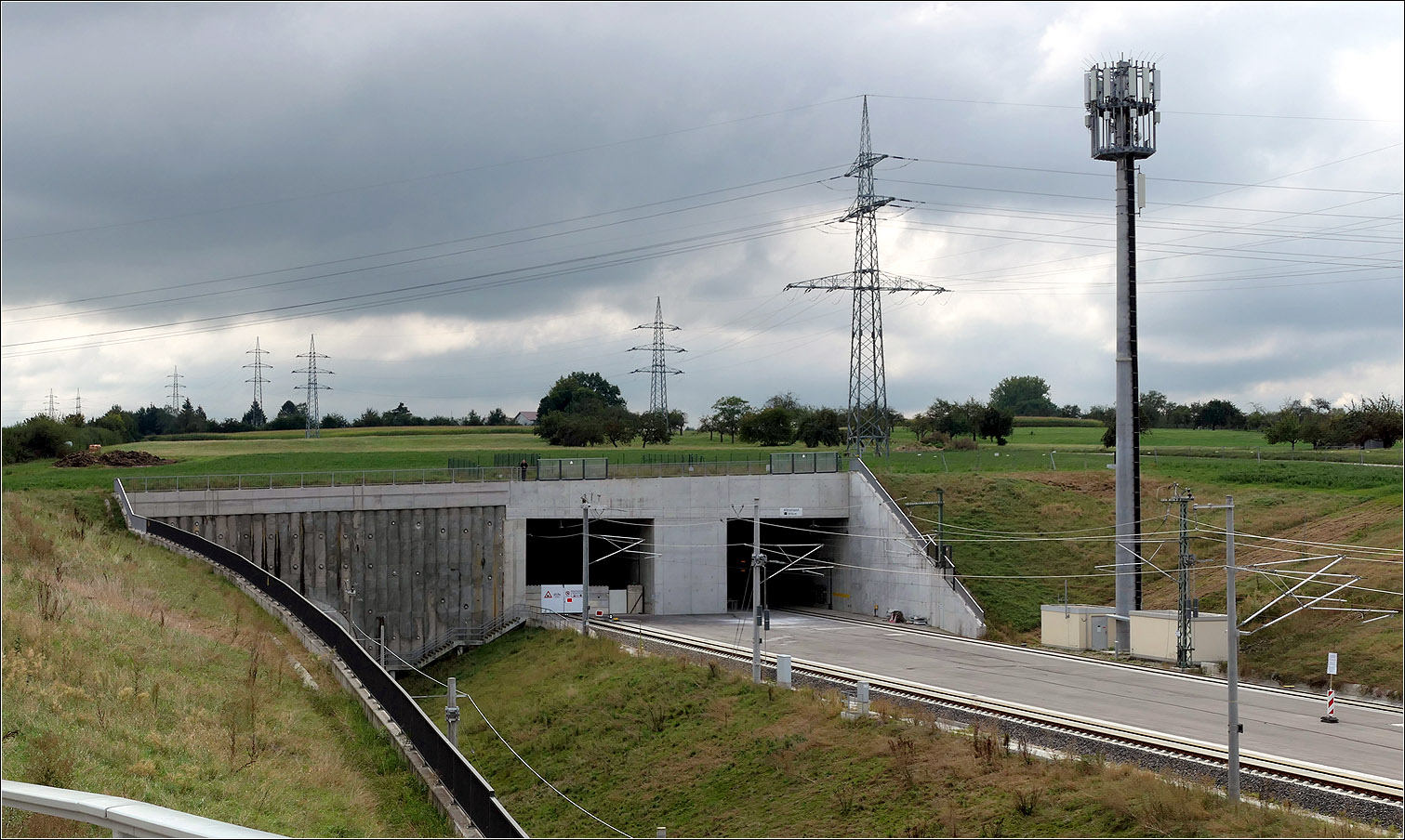 Eckige Tunneleinfahrten - 

Westportal des 8174 m langen Albvorlandtunnels bei Wendlingen. Links im Bild ist die tiefer liegende Güterzuganbindung zu erahnen. Über diese eingleisige Verbindungskurve rollt heute der gesamte Bahnverkehr der seit Ende 2022 auf der Neubaustrecke rollt. Da der erste Gleiswechsel erst auf der anderen Seite des Tunnels liegt, muss derzeit noch auf einen recht langen Abschnitt eingleisig gefahren werden, was die Leistungsfähigkeit des fertigen Abschnittes der Neubaustrecke noch einschränkt. Daher fahren auch noch einige ICEs über die Altstrecke und die Geislinger Steige.

Im Albvorlandtunnel vereinigt sich das Gleis der Güterzuganbindung mit dem Gleis der nördlichen Tunnelröhre. 

13.09.2024 (M)