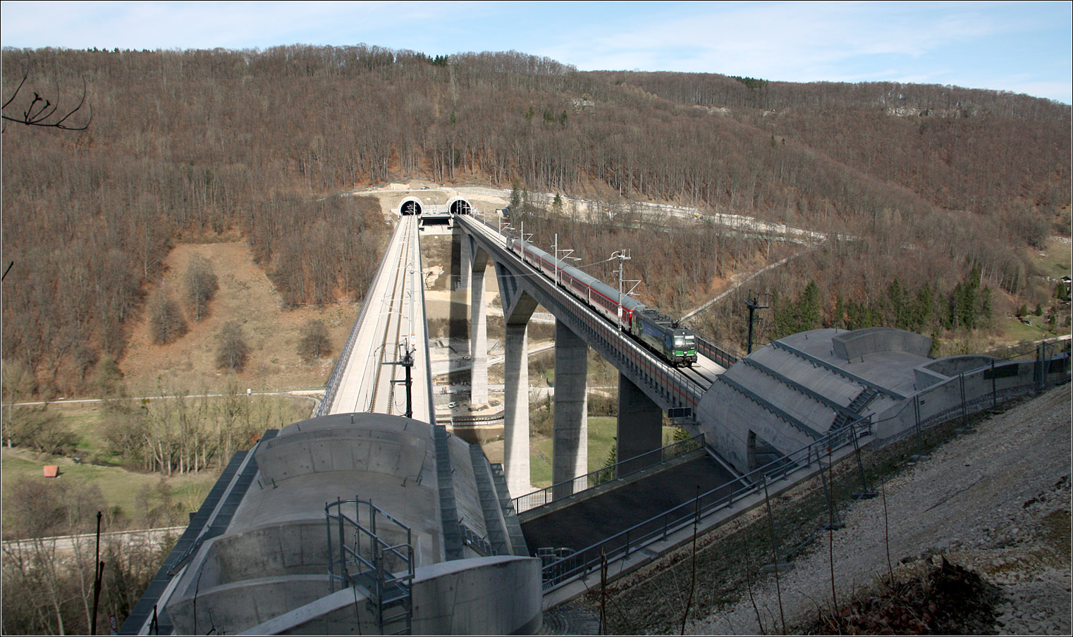 Hoch über dem Filstal - 

Am unteren dem westliche Portal des Steinbühltunnels kommt die Schnellfahrstrecke für kurz aus dem Berg um das Filstal in 85 m Höhe zu queren. Danach geht es wieder in den Berg um nach 8,8 km am Aichelberg wieder ins Tageslicht zu kommen. Die zwei Röhren des Boßlertunnels wurden mit Tunnelbohrmaschinen aufgefahren.

Hier quert ein IRE 200 in Fahrtrichtung Wendligen das Tal.

17.03.2022 (M)