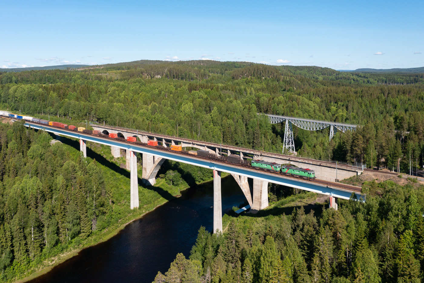 Kurz nach dem ARE nach Narvik folgt am 17.06.2022 mit rund 100 Minuten Verspätung der 4302 mit einer Doppeltraktion Green Cargo Rc4 über die Tallbergsbroarna bei Nyåker. Schön zu sehen sind die drei Brücken. Die hinterste und älteste Brücke ist die Straßenbrücke von 1891. Die mittlere Brücke ist die erste Eisenbahnbrücke und stammt aus dem Jahr 1919. Die vorderste Brücke ist die jüngste und hat 1994 die alte Eisenbahnbrücke ersetzt.