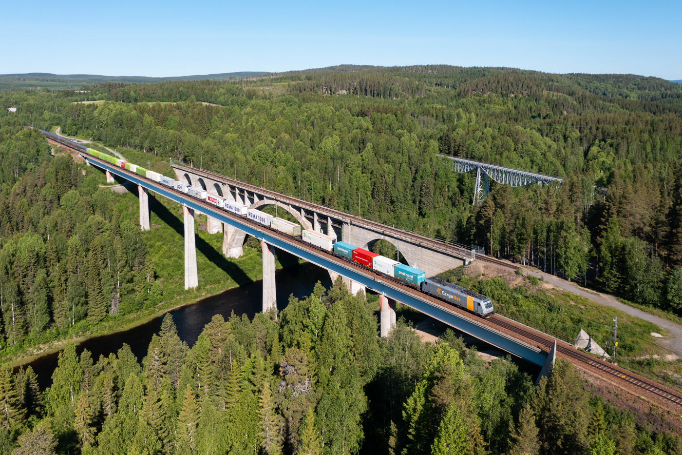 Mitten im schwedischen Wald liegen die Tallbergsbroarna, drei Brücken über den Öreälven, nahe der Gemeinde Nyåker südwestlich von Umeå. Leider ist an der Stambanan genom övre Norrland die Vegetation oft so dicht, dass es unmöglich ist gute Fotostandorte zu finden. Daher bleibt oft nur die Möglichkeit den fliegenden Helfer einzusetzen. Morgens verkehren mehrere Güterzüge in dichter Folge in Richtung Norden. Da die gesamte Strecke eingleisig ist wird oft in diesen Kolonnen gefahren. Den Anfang macht am 17.06.2022 der erste der beiden Arctic Rail Express Züge (41916) nach Narvik.