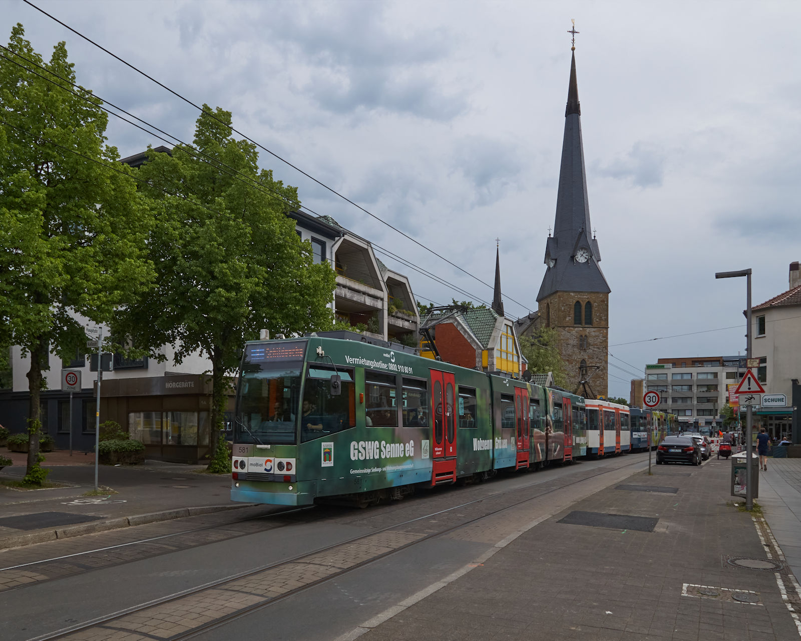 Mittlerweile wird die Strecke in der Brackweder Hauptsraße in Bielefeld auf Stadtbahnniveau umgebaut. Als M8D 581 mit seinem Zug aus einem weiteren M8D und einem MB4 am 16.05.2023, nahe der Brackweder Kirche auf dem Weg nach Schildesche war, war davon noch nicht viel zu sehen.  