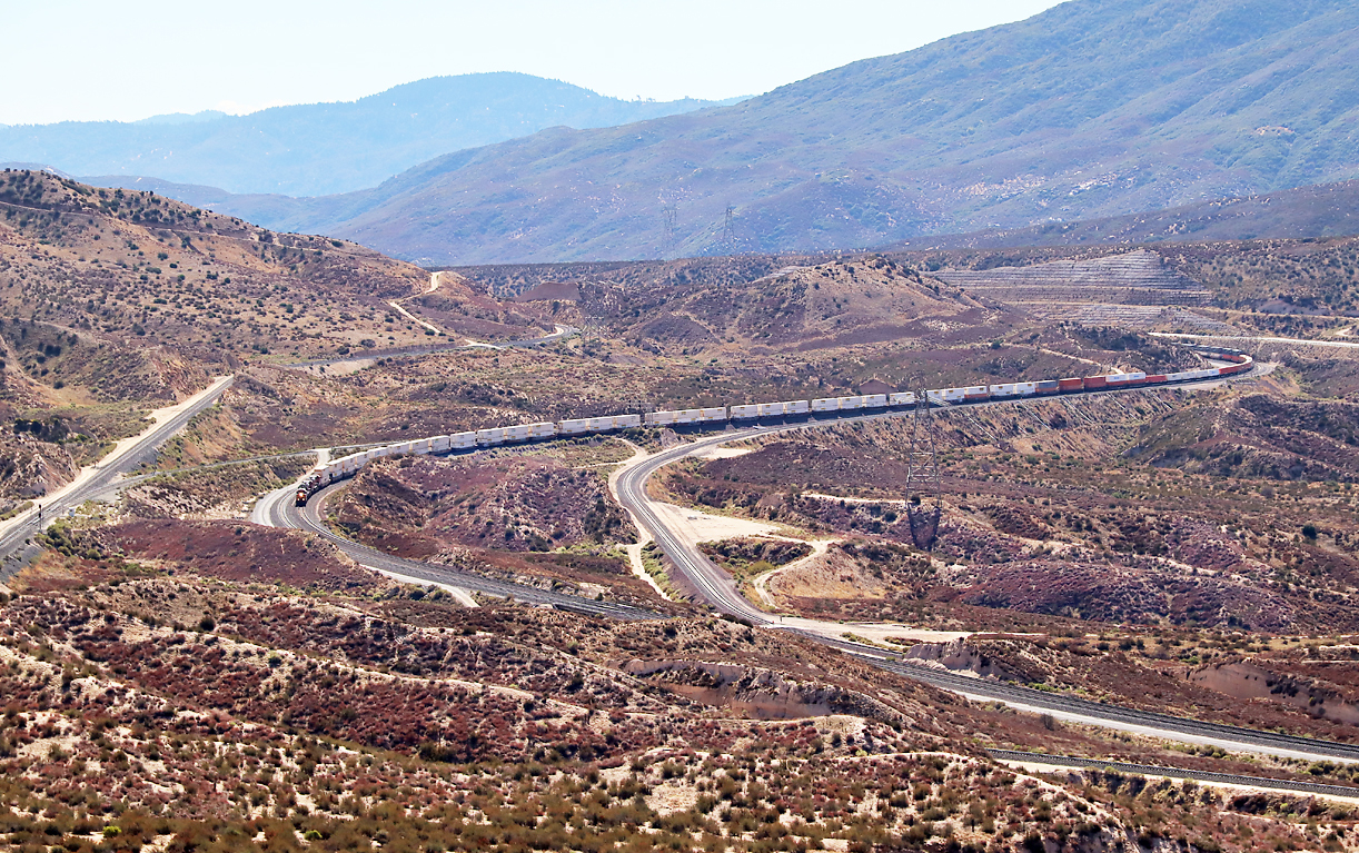 Modelleisenbahn? Nein, sehr spektakuläre Linienführung am Cajon Pass: Uebersicht über den südlichen, obersten Teil des Cajon Passes, aufgenommen von einem Parkplatz vom Interstate-Highway: die Passhöhe (Summit) für die Eisenbahn und die Hauptstrasse befindet sich im Hintergrund hinter dem Hügel etwas rechts von der Bildmitte. Links das Gleis der Union Pacific (UP), an dieser Stelle einspurig, weiter unten doppelspurig. Der Containerzug fährt auf der 'mittleren Linie' (durchgehend doppelspurig) talwärts, diese Gleise gehören der BNSF. Auf diesen beiden BNSF-Gleisen wird berg- und talwärts gefahren. Links der Lokomotiven ist ein Verbindungsgleis vom UP- zum BNSF-Gleis zu sehen. Das Gleis rechts (durchgehend einspurig) gehört ebenfalls der BNSF und wird grundsätzlich nur talwärts befahren, da dieses Gleis ein stärkeres Gefälle aufweist. Vereinzelte, leichte Züge (zB der Amtrak-Southwest-Chief Los Angeles - Chicago oder Lokzüge) nützen dieses Gleis auch für die Bergfahrt. Ganz rechts hinter dem hintersten Zugteil ist die Hauptstrasse zu sehen, auf dieser sind mit dem Auto sehr gute Fotostellen beim 'Summit' ziemlich leicht zu erreichen. Rechts im Anschluss von diesem Bild liegt der bei Eisenbahn-Fotografen bekannte Fotopunkt 'Hill 582' in der Nähe der mittleren Doppelspur (Fotos folgen). Cajon Pass, CA, 24.9.2022
